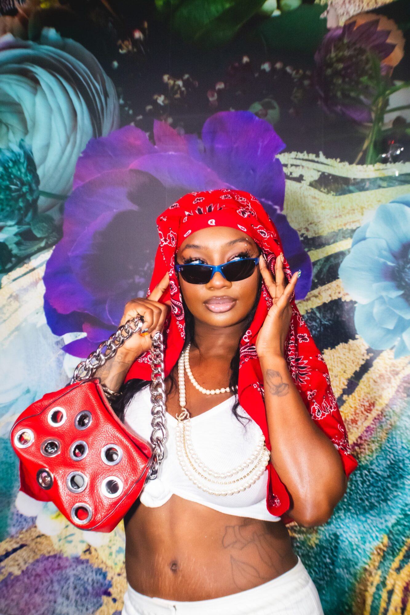 Woman with sunglasses, red headscarf, white crop top, pearl necklace, holding a red bag, standing in front of colorful floral background.