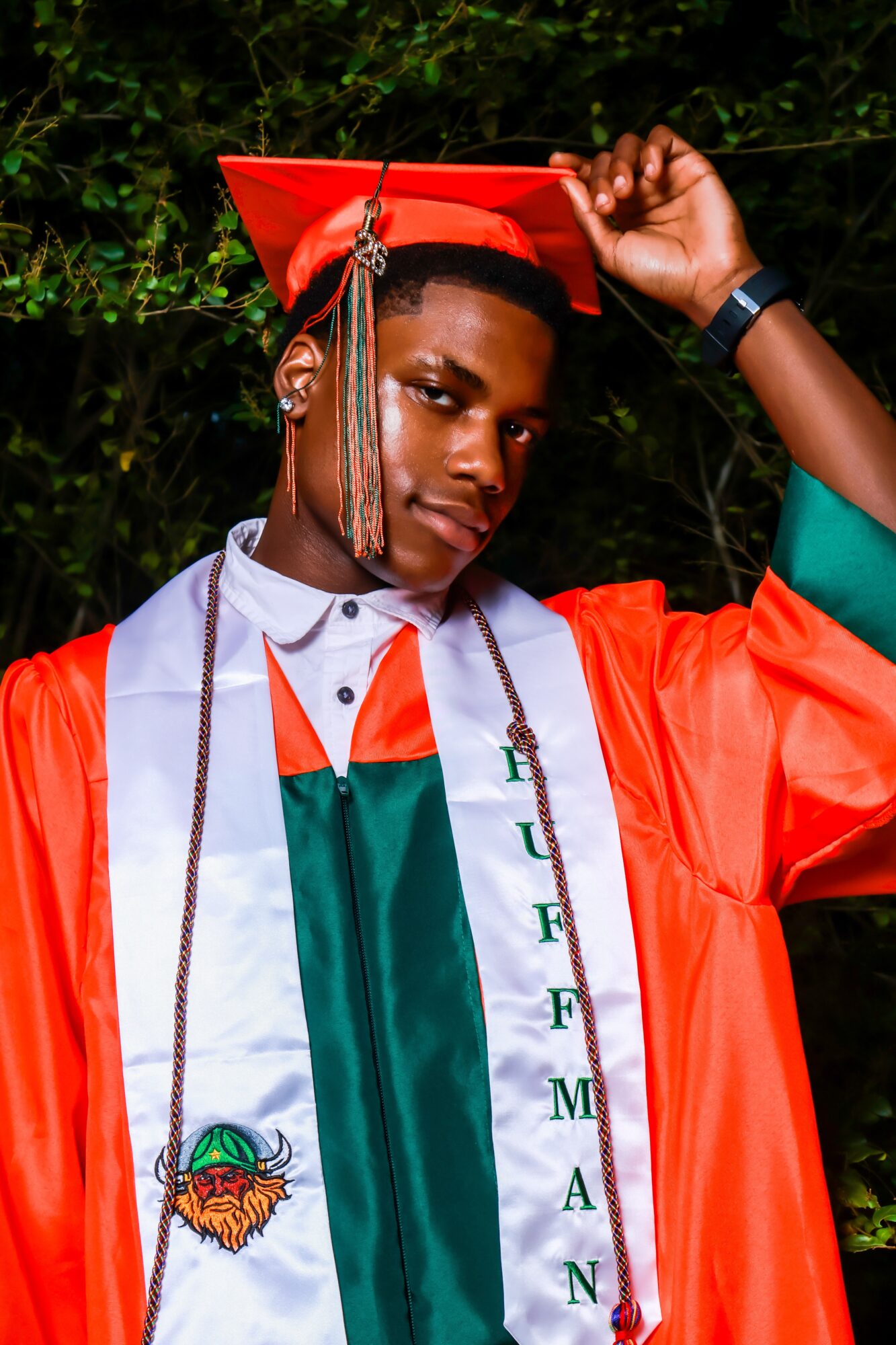 Young man in graduation cap and gown, adjusting cap, outdoors with greenery background