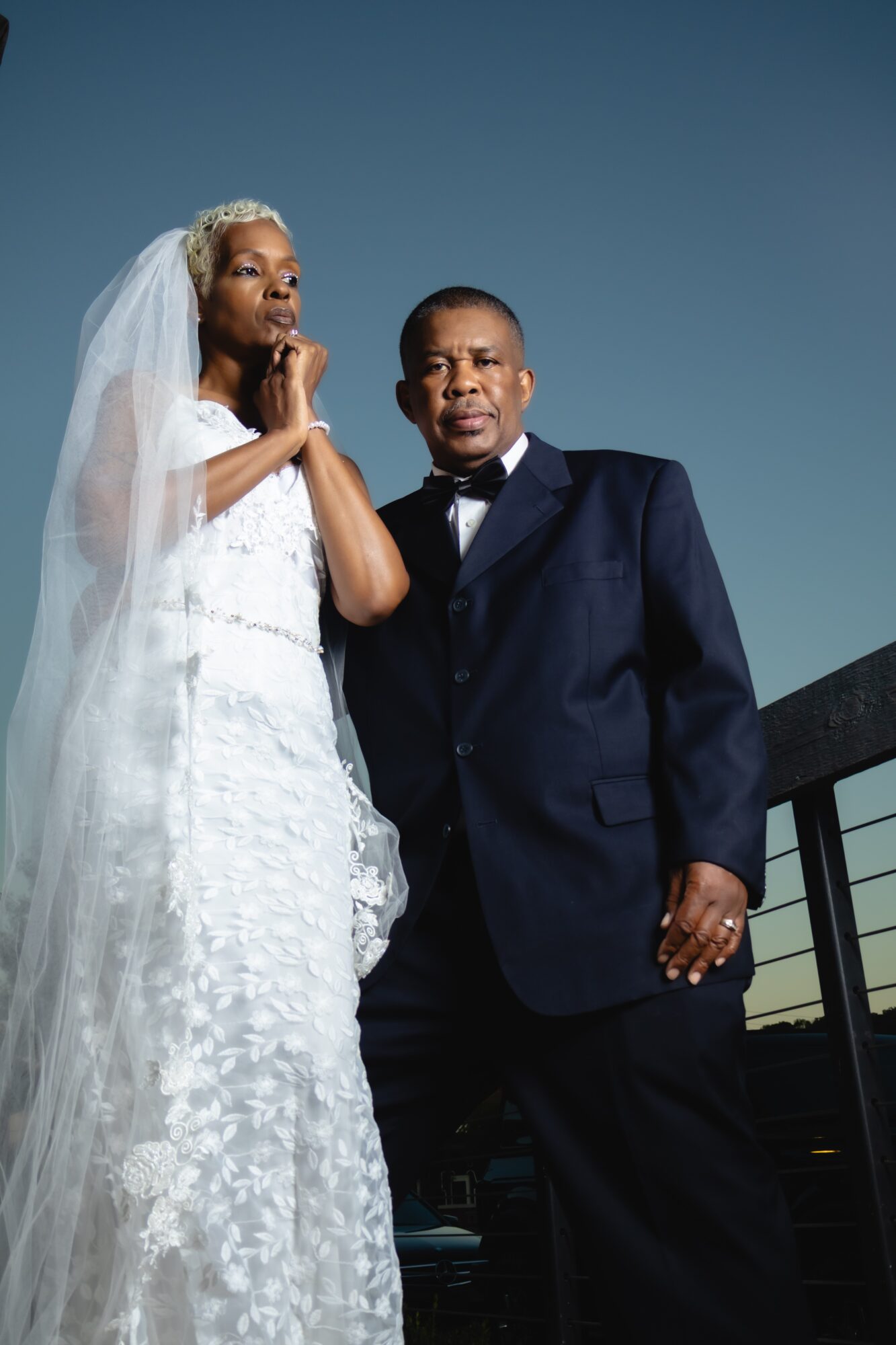 A bride and groom stand together outdoors against a darkening sky, dressed in wedding attire, with the bride wearing a veil and gown, and the groom in a suit.
