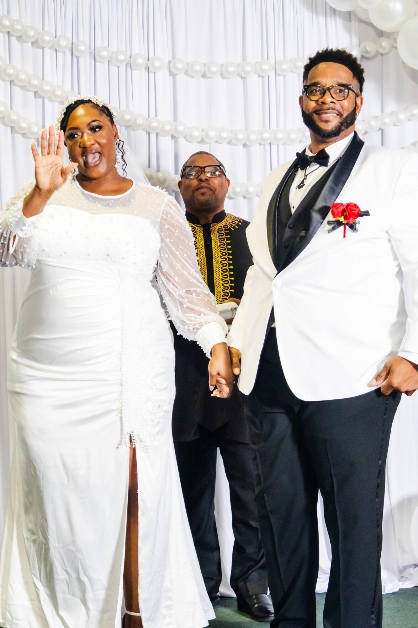 A couple holding hands at a wedding, with a person in the background, all smiling, in front of a decorated backdrop.