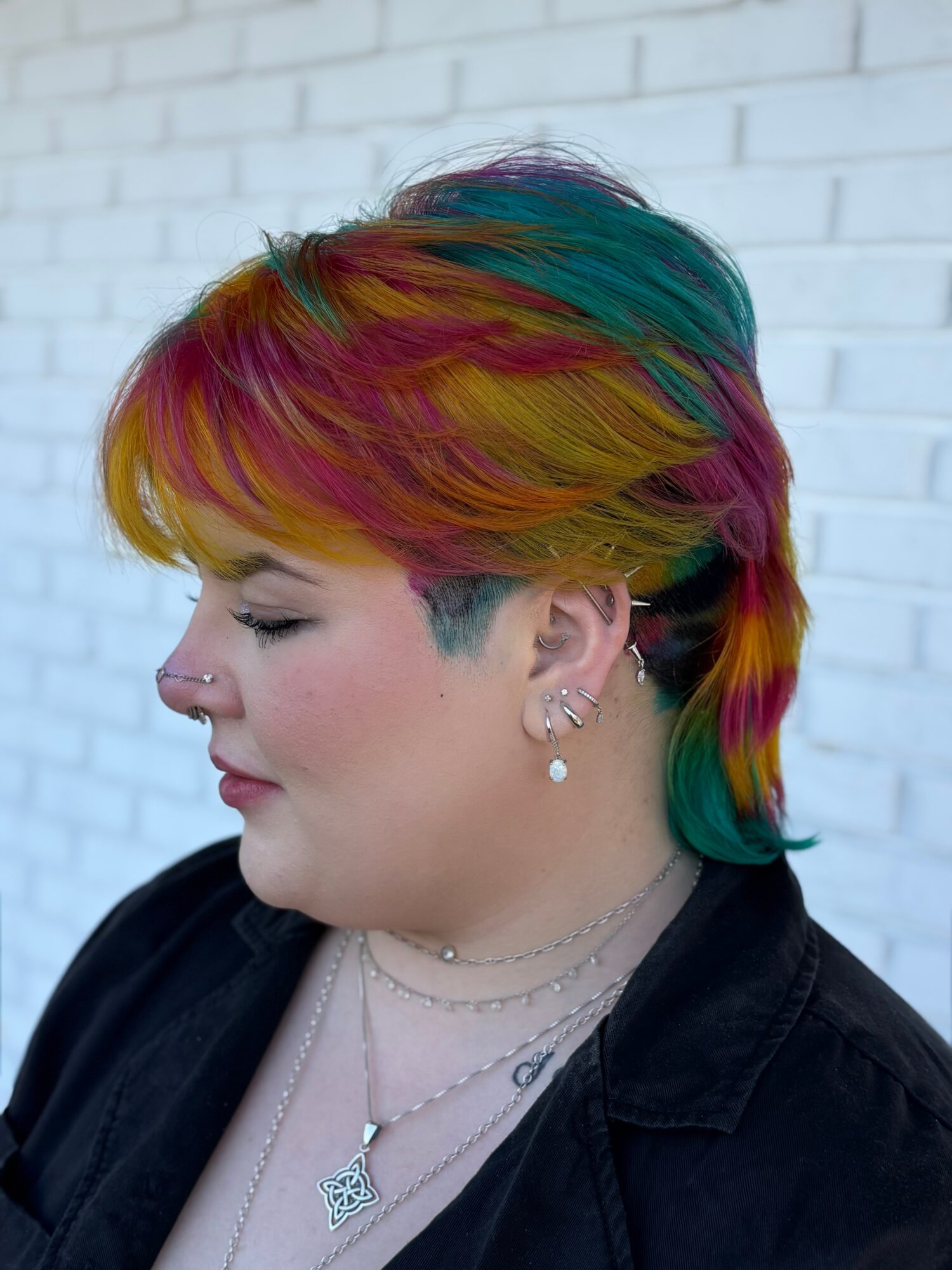 Side profile of a person with multicolored short hair, wearing earrings and layered necklaces, against a white brick wall.