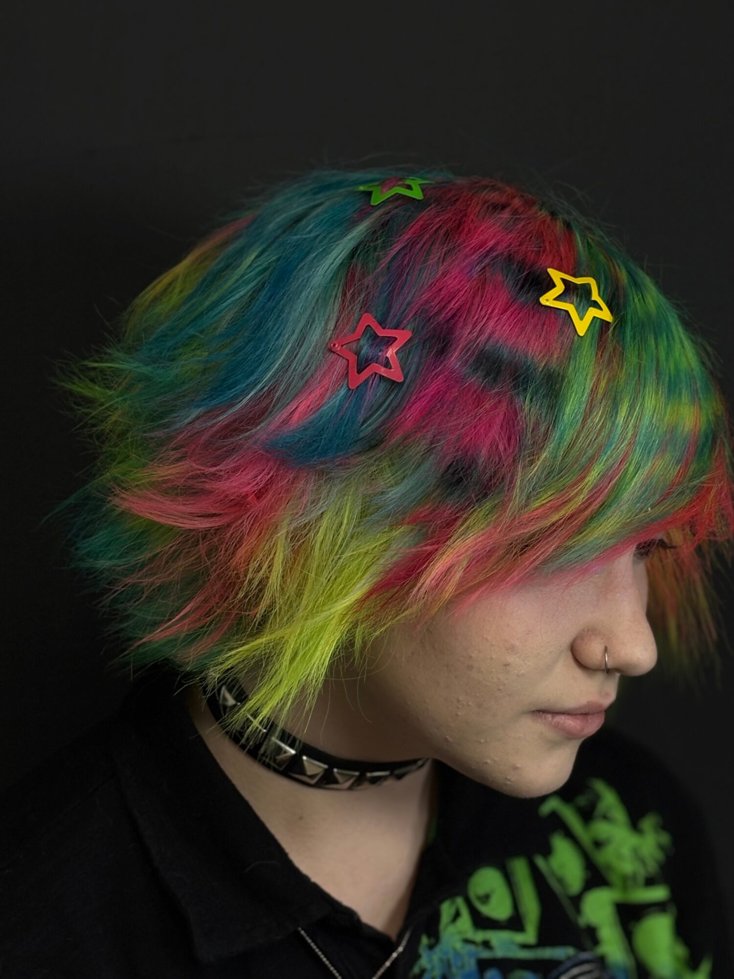 Person with multicolored hair and star-shaped hair clips, wearing a choker and black shirt, against a dark background.