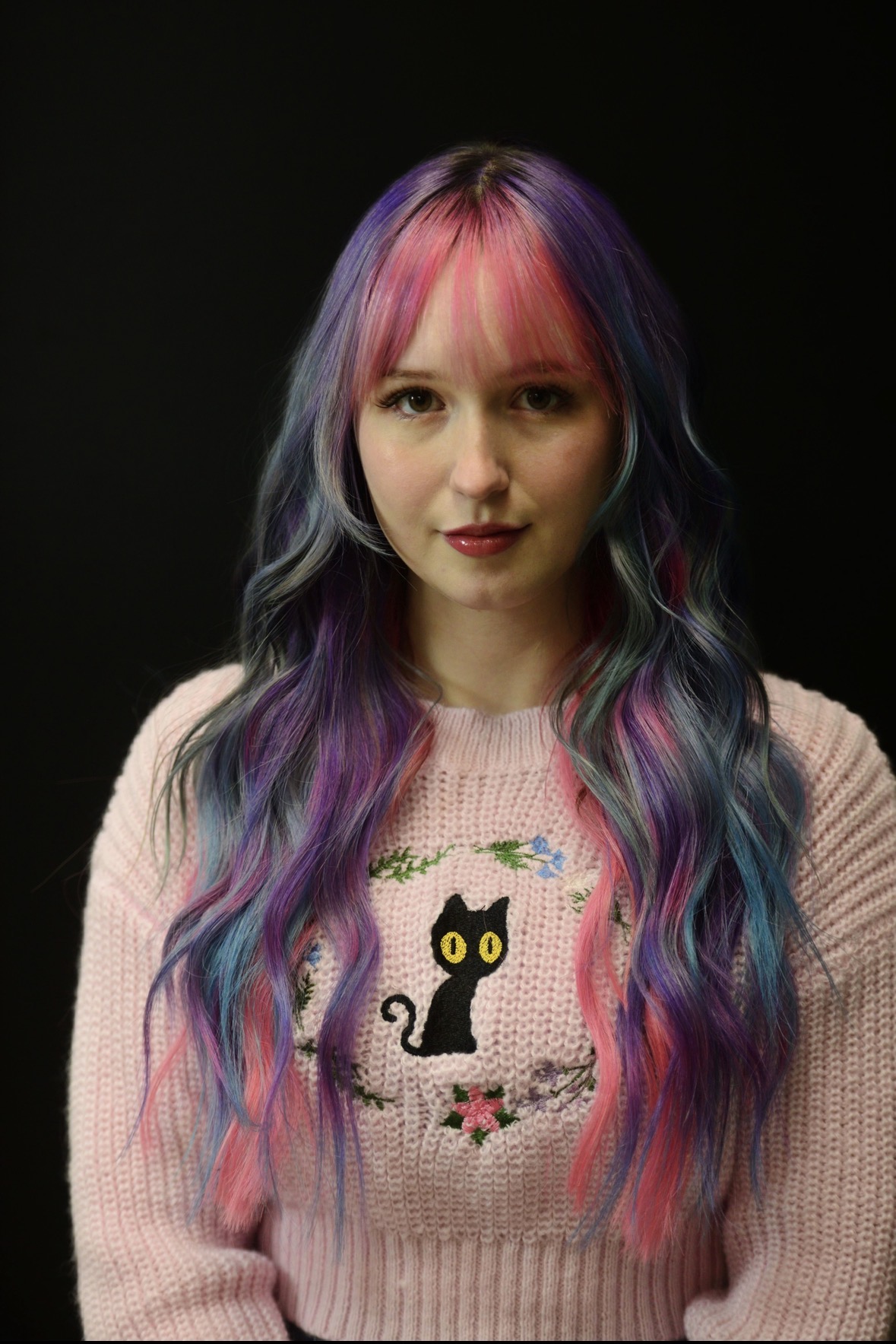 Young woman with multicolored hair, wearing a pink sweater with a black cat and floral design, against a black background.