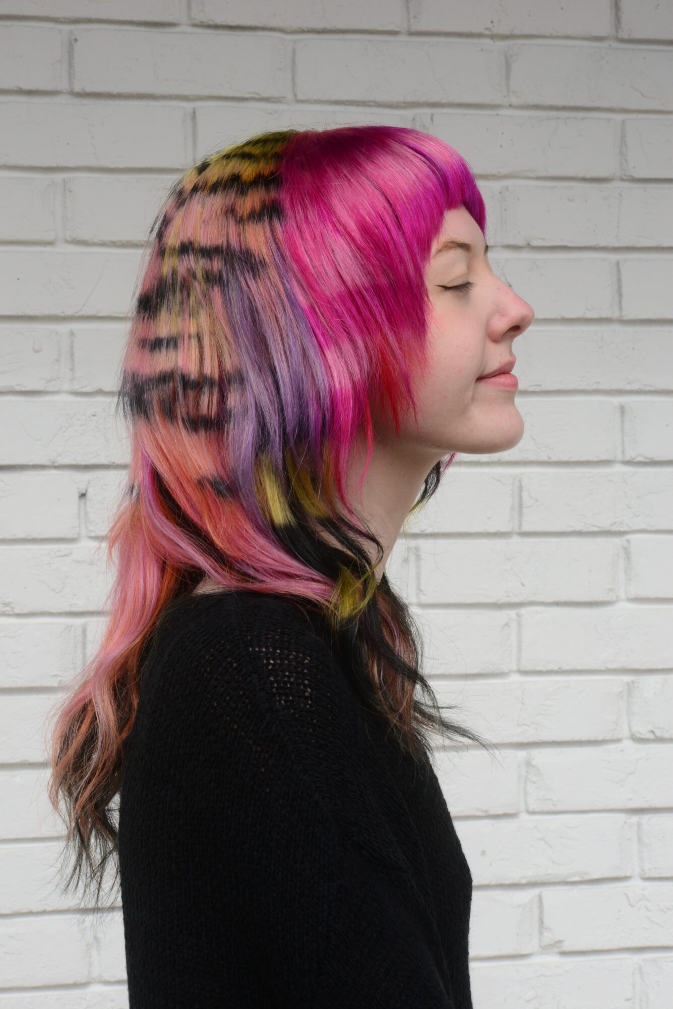 Side profile of a woman with colorful, multi-hued hair against a white brick wall.