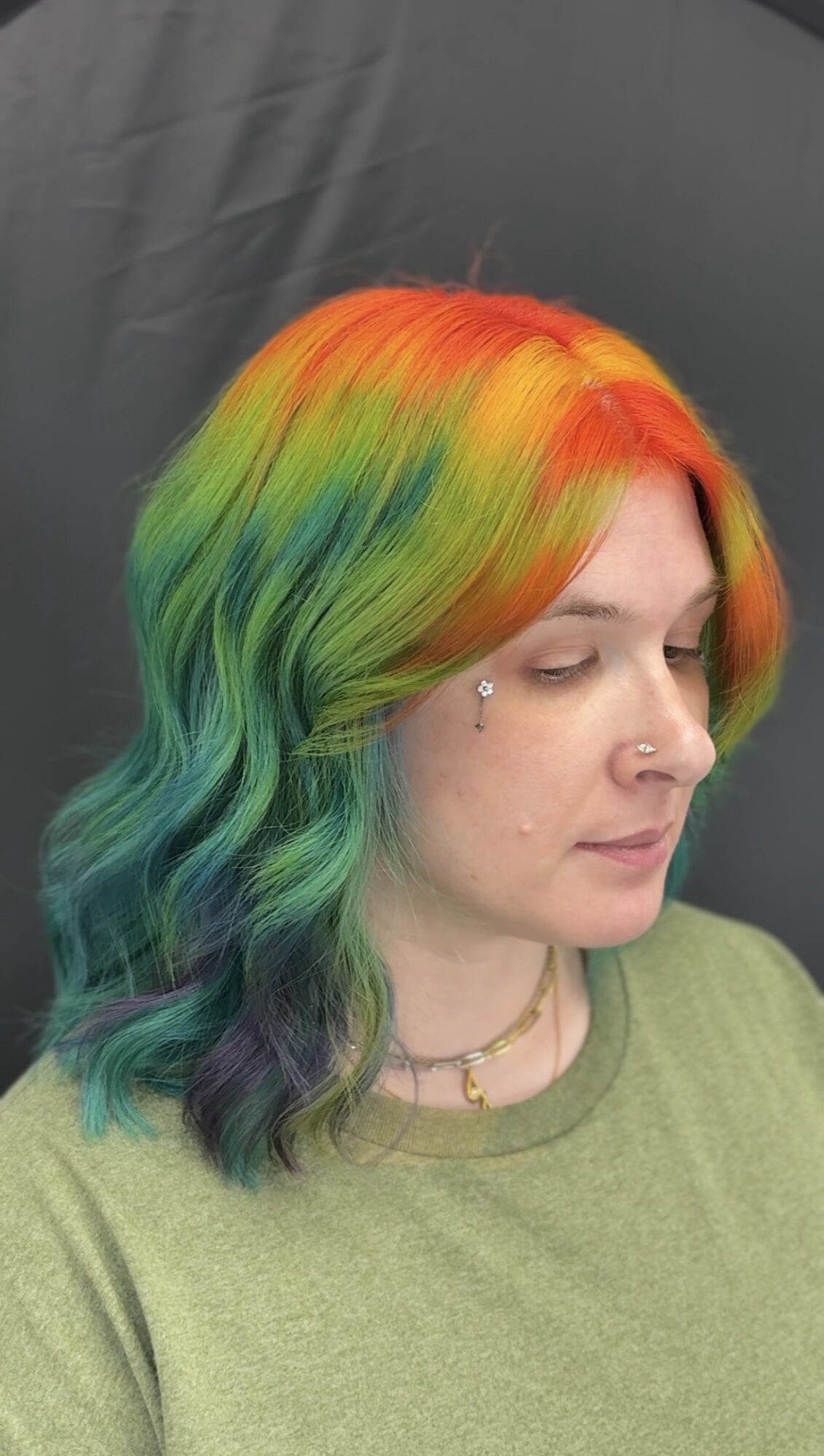 Woman with rainbow-colored hair, looking down, wearing a green shirt and a necklace, against a dark background.