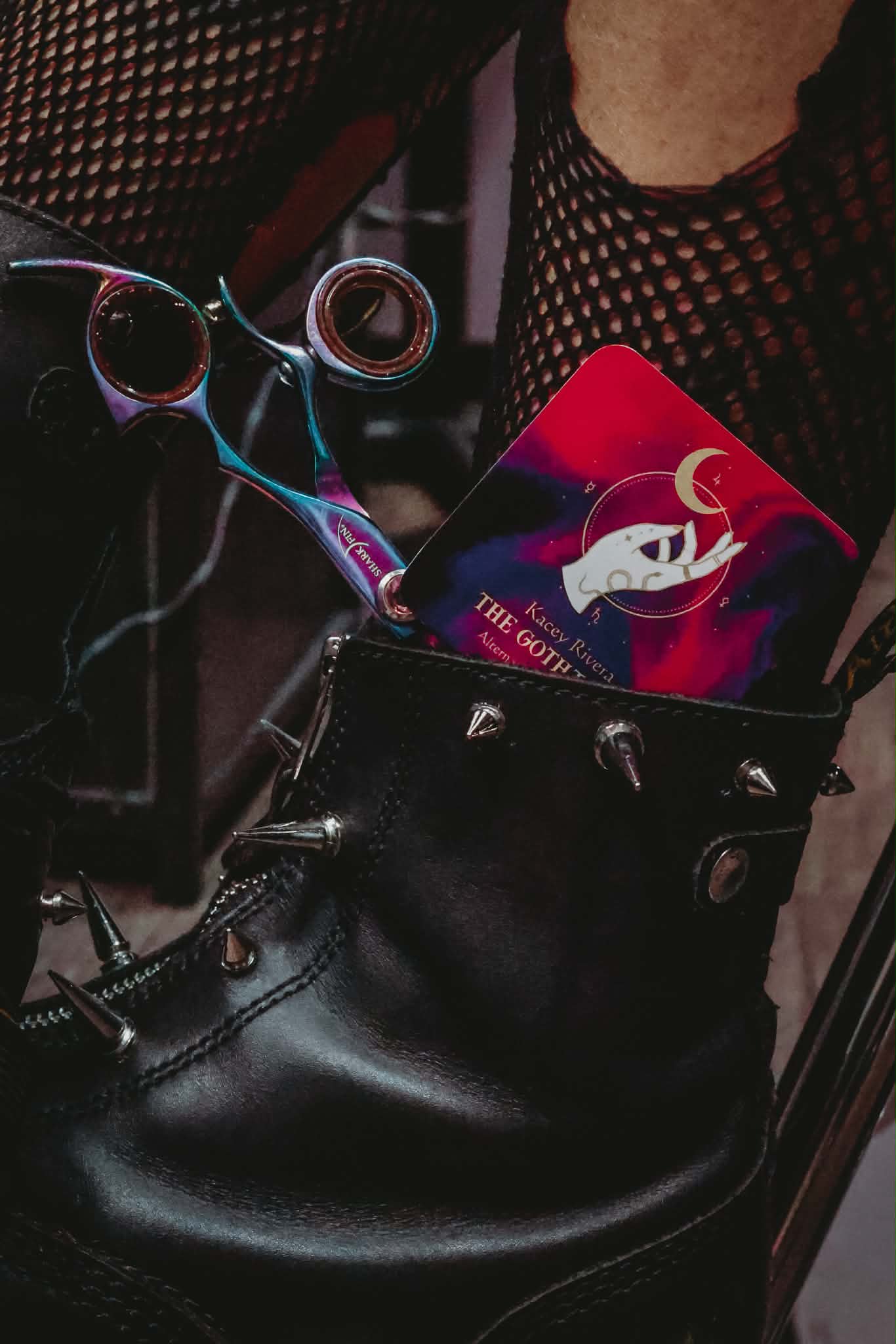Black leather bag with a pink and purple card inside, sunglasses hanging on the bag, and a person wearing a black fishnet top.