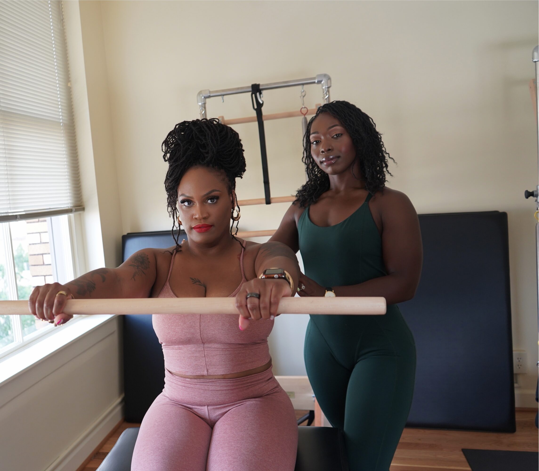 Two women in workout attire in a fitness studio, one sitting and holding a ballet barre, the other standing behind her.