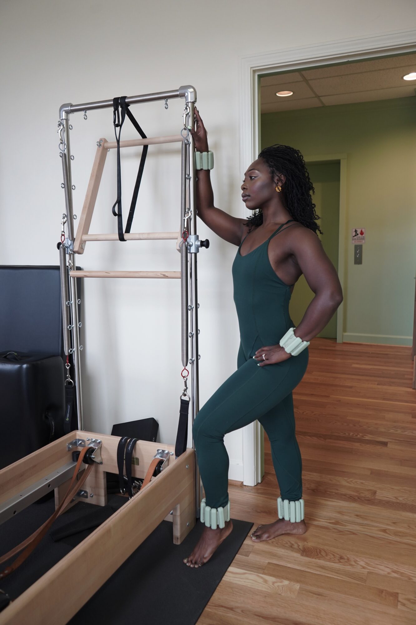 Woman in green workout outfit standing next to Pilates equipment in a room with wooden floor and green walls.