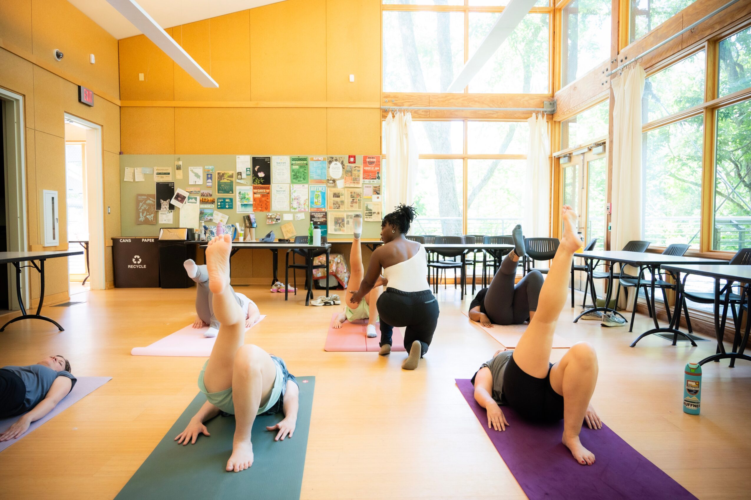 People practicing yoga in a bright, spacious room with large windows and a bulletin board on the wall.
