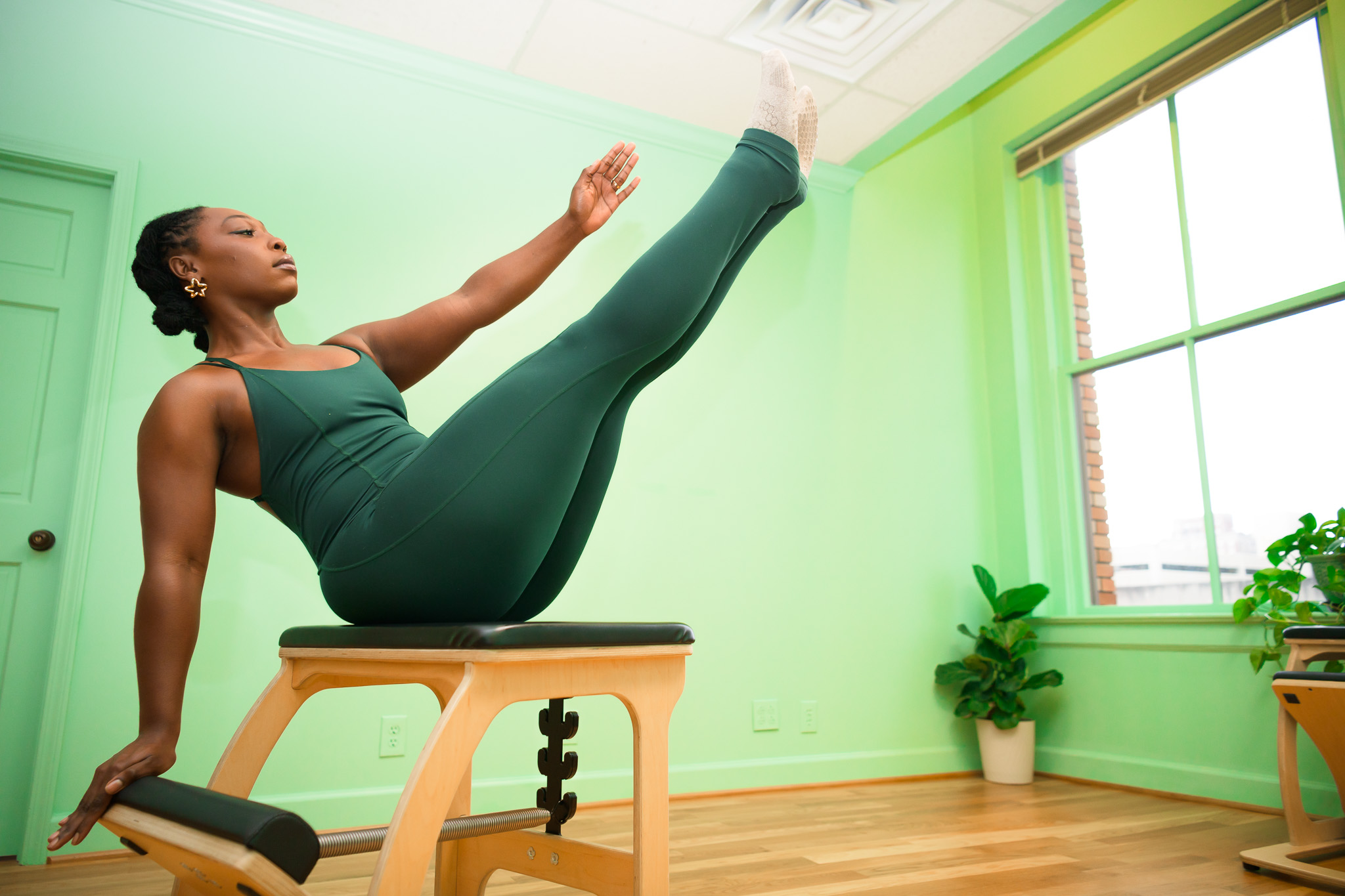 Woman practicing yoga on a chair in a bright room with green walls and large window, balancing with one leg raised.