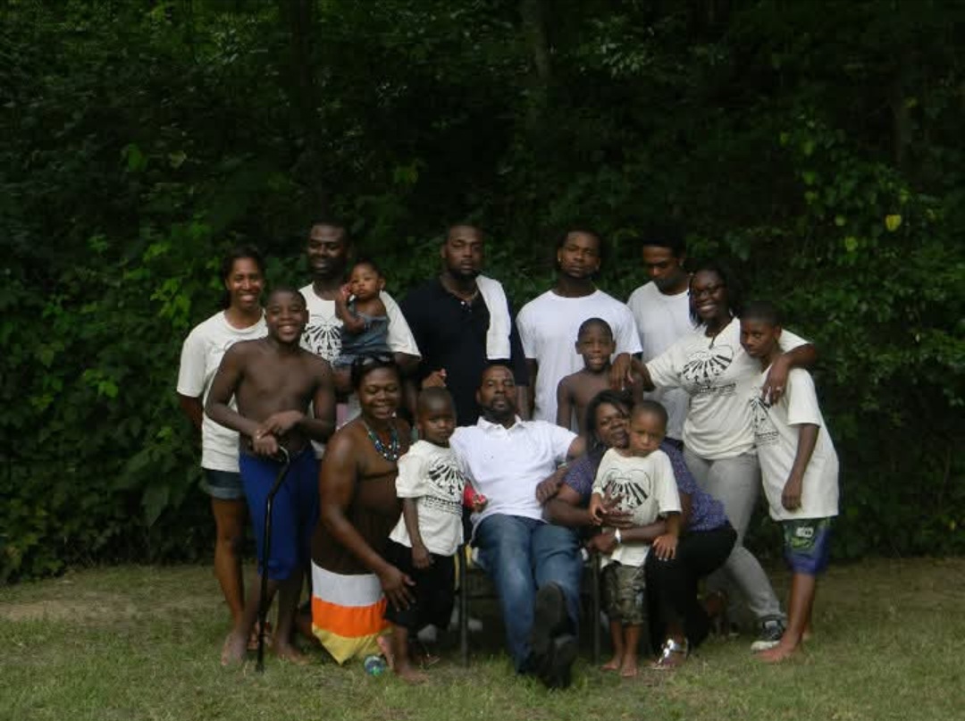Group of people outdoors in front of trees, some standing and some sitting, smiling at the camera.