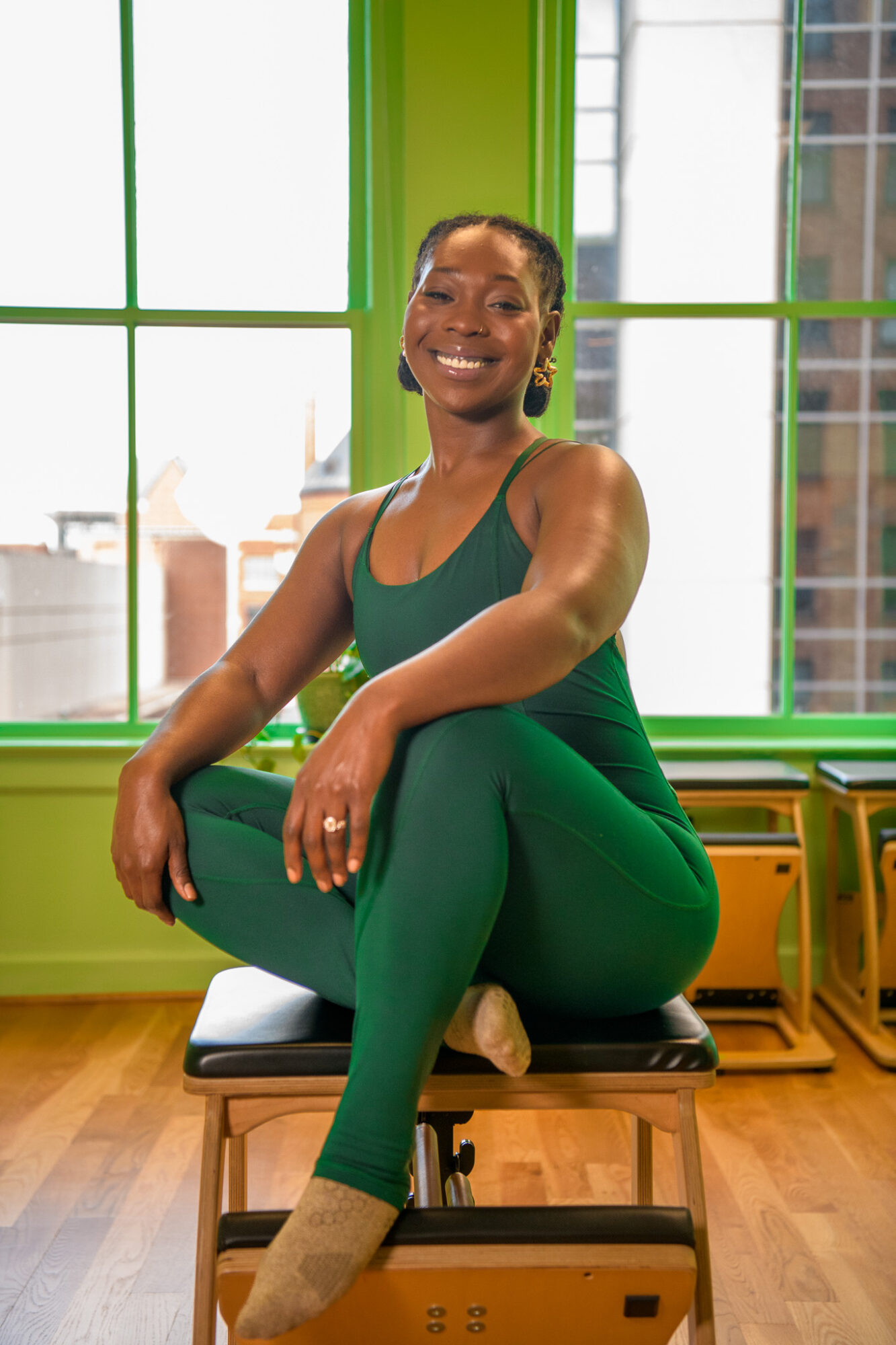 Smiling woman in green workout clothes sitting on a bench indoors with large windows and cityscape view behind her.