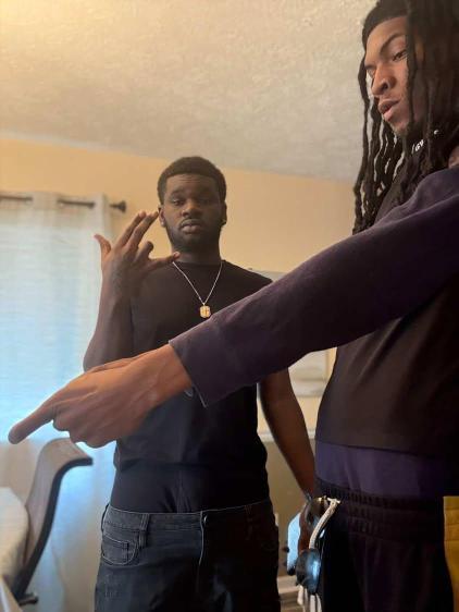 Two young men indoors, one with dreadlocks and the other with short hair, gesturing with their hands. The room has a window with curtains.