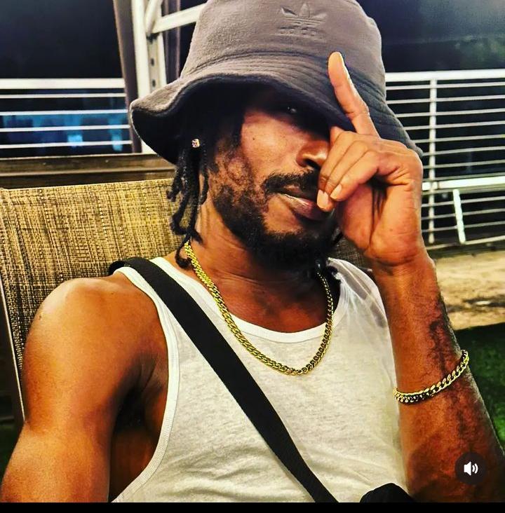 Man wearing a bucket hat, sleeveless shirt, and gold chain, sitting outdoors with hand near face, looking at camera.