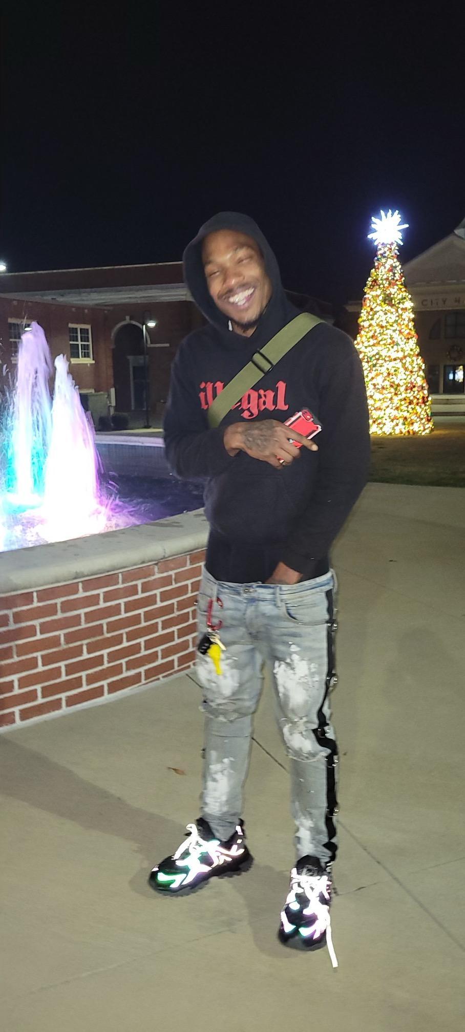 Young person smiling, wearing a black hoodie and ripped jeans, standing outdoors at night with festive lights and a fountain.