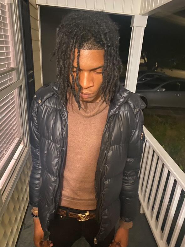 Young man with dreadlocks wearing a black jacket and brown shirt, standing on a porch at night.