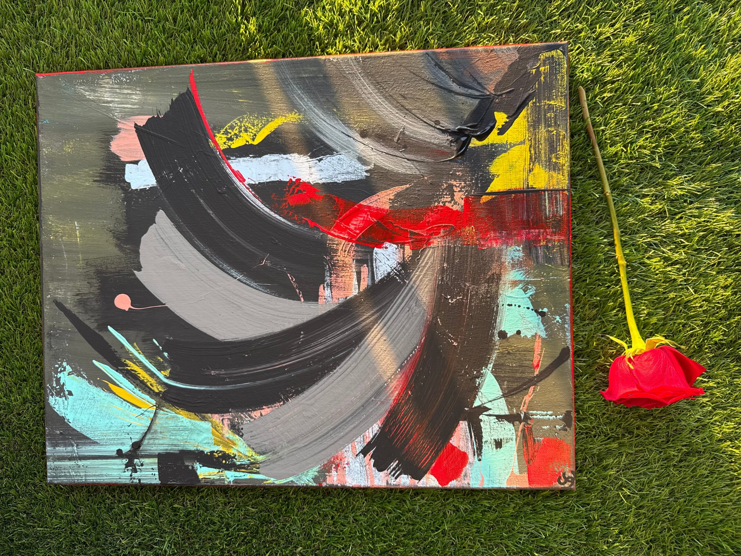 Abstract painting with black, red, yellow, and light blue brushstrokes on a canvas, placed on grass with a yellow stick and red flower nearby.