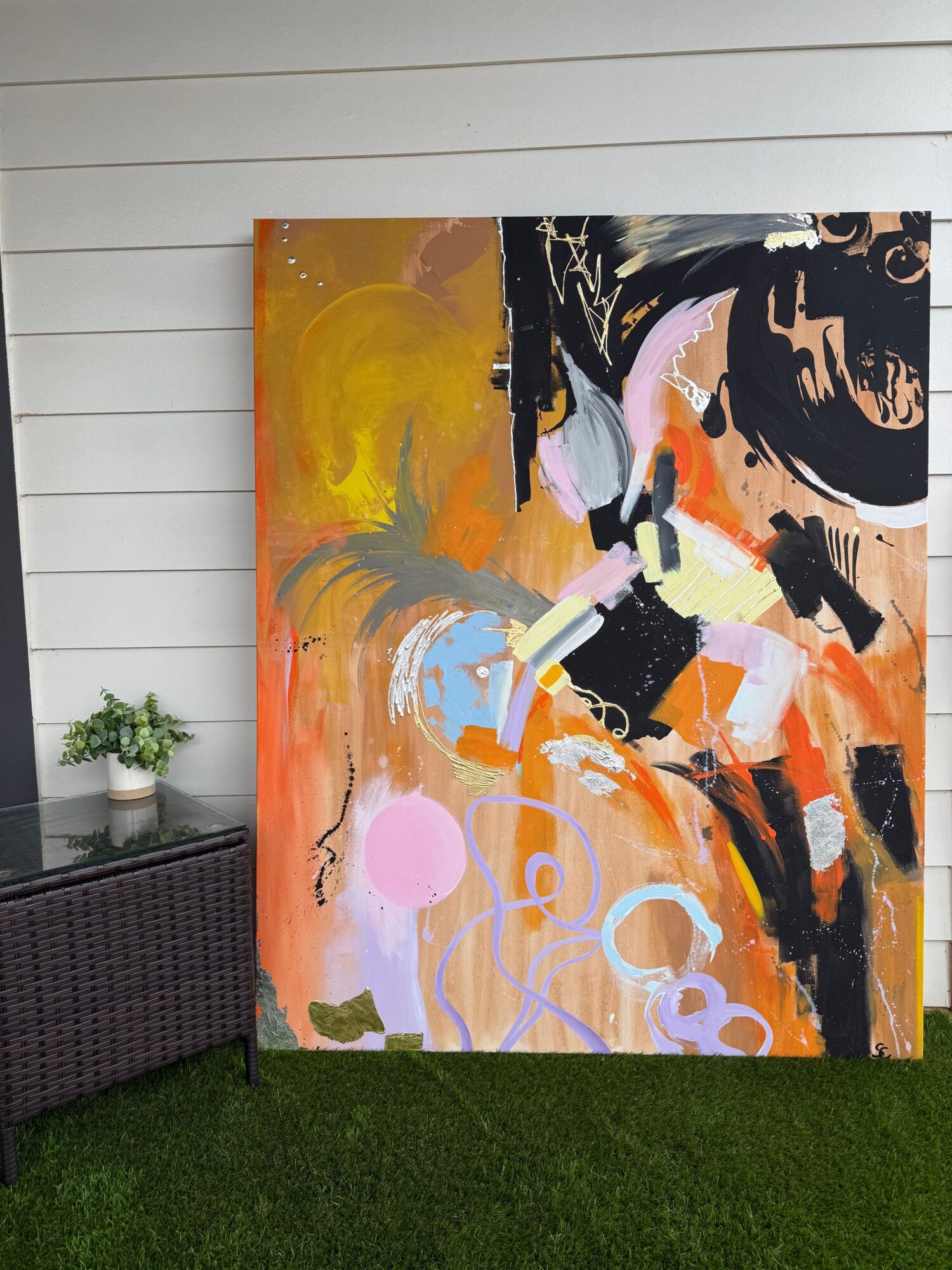 Abstract painting with black, orange, pink, and purple brushstrokes on a canvas, leaning against a white wall on grass.