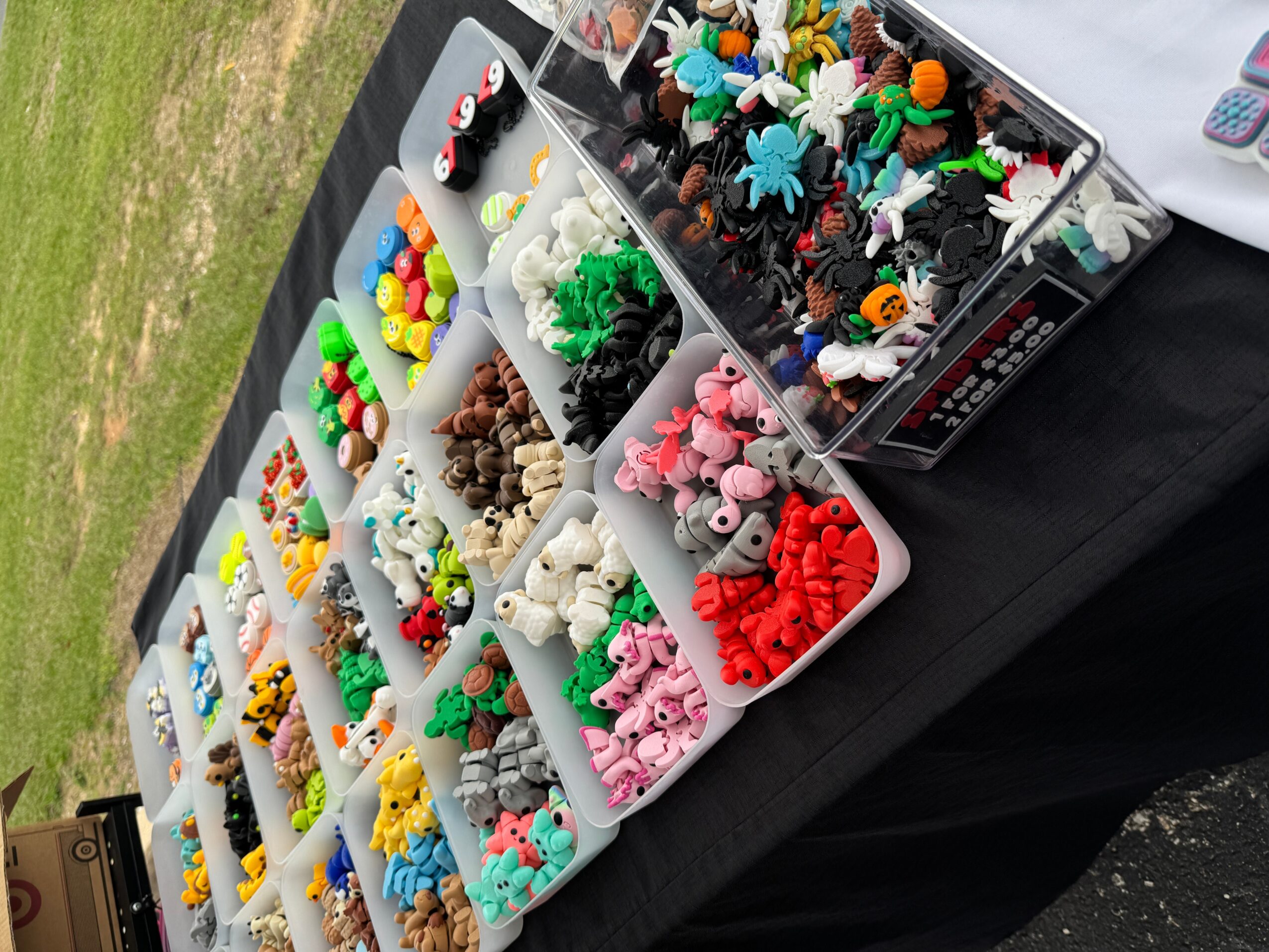 Display of colorful small toys and figurines in plastic containers on a black table outdoors.