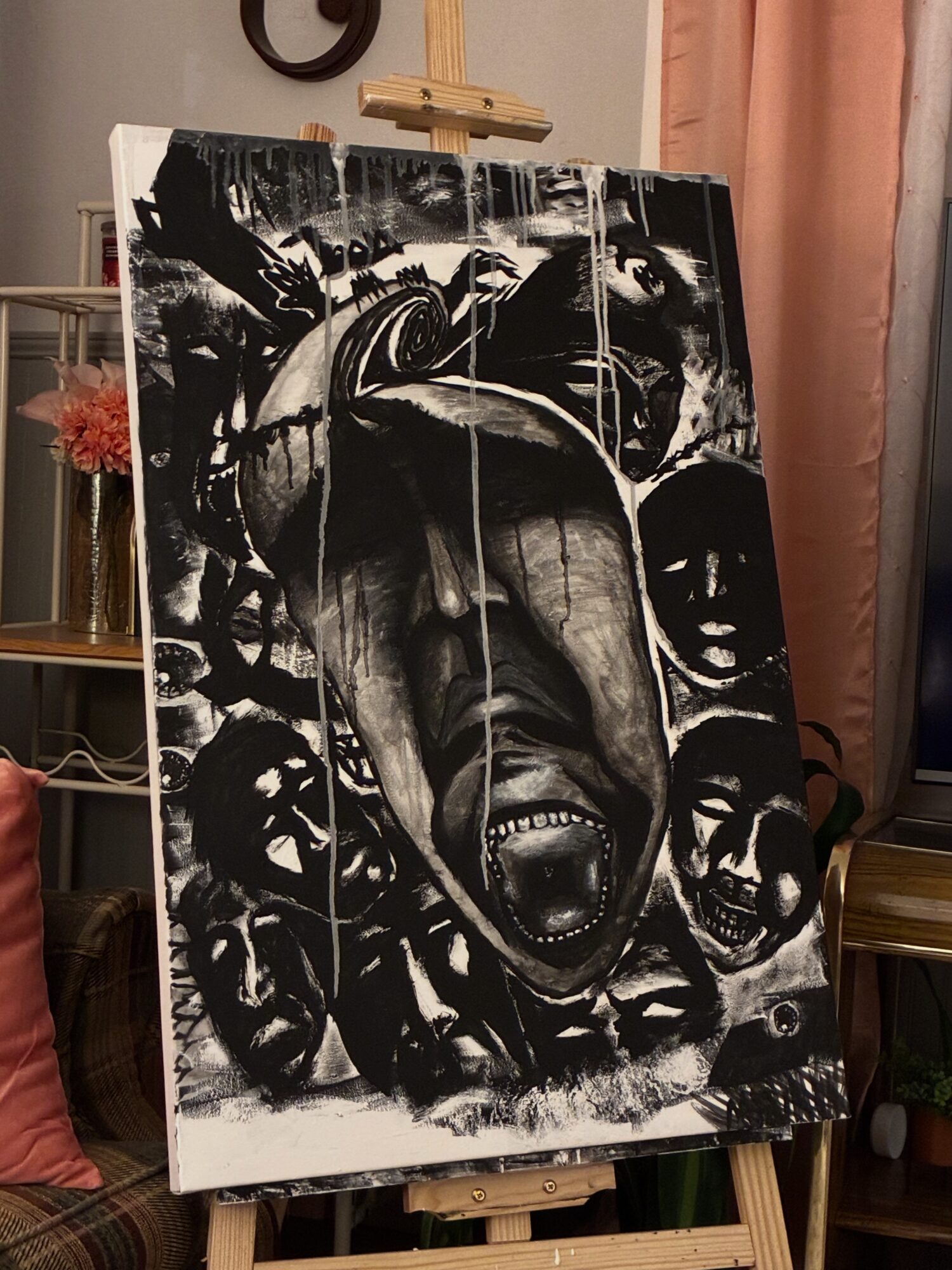 Black and white painting of a screaming face surrounded by multiple smaller faces, on an easel indoors.