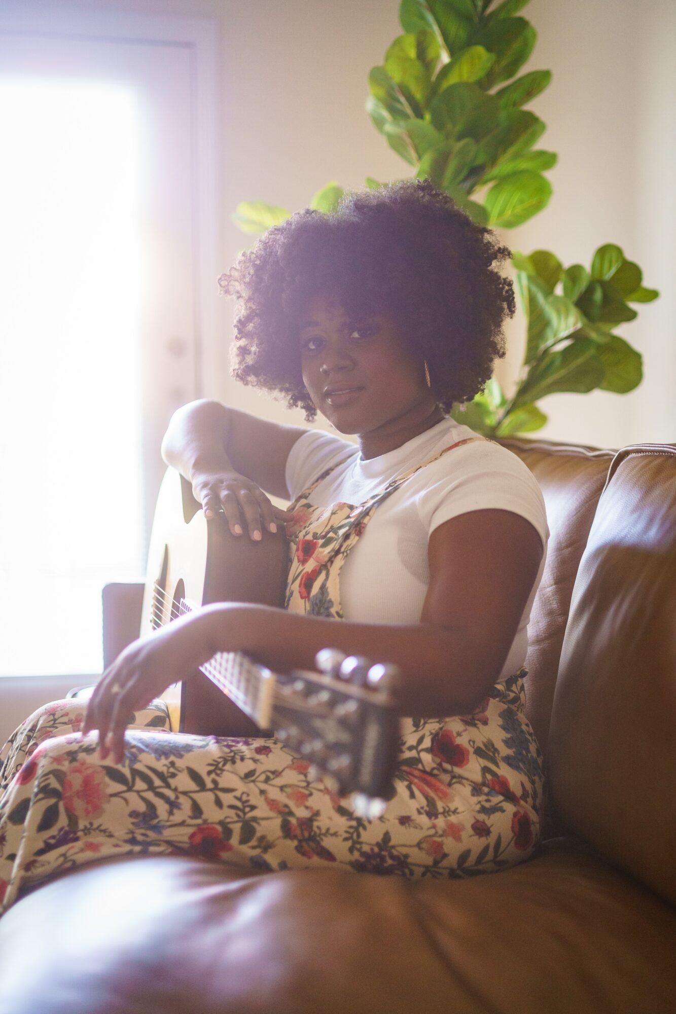 Young woman with curly hair sitting on a leather couch, with a large green plant behind her, sunlight streaming in.
