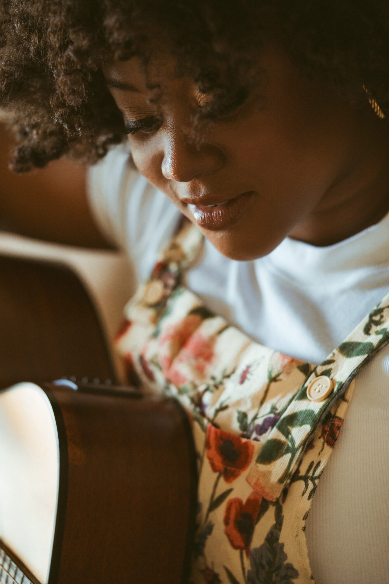 Close-up of a woman with curly hair looking down, wearing a floral dress and white top, playing a guitar.