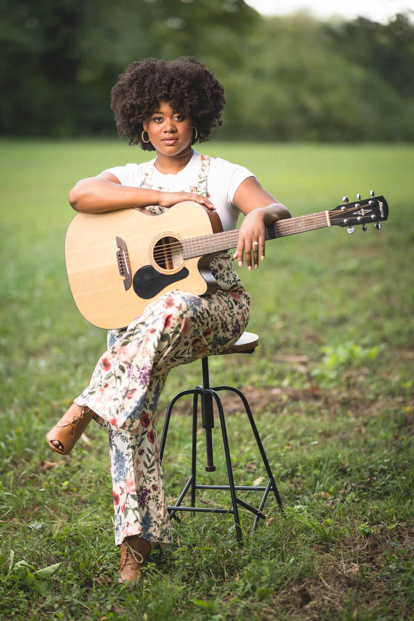 Young girl with curly hair sitting on a stool outdoors, holding an acoustic guitar, in a grassy field with trees in background.