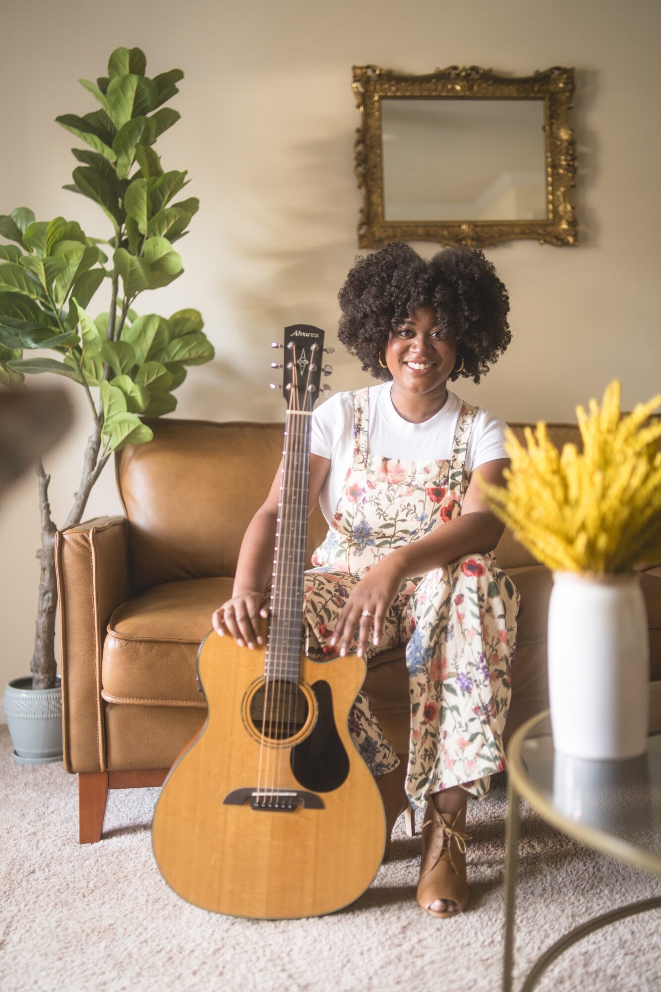Woman with curly hair sitting on a sofa, holding a guitar, in a living room with a plant and a mirror.