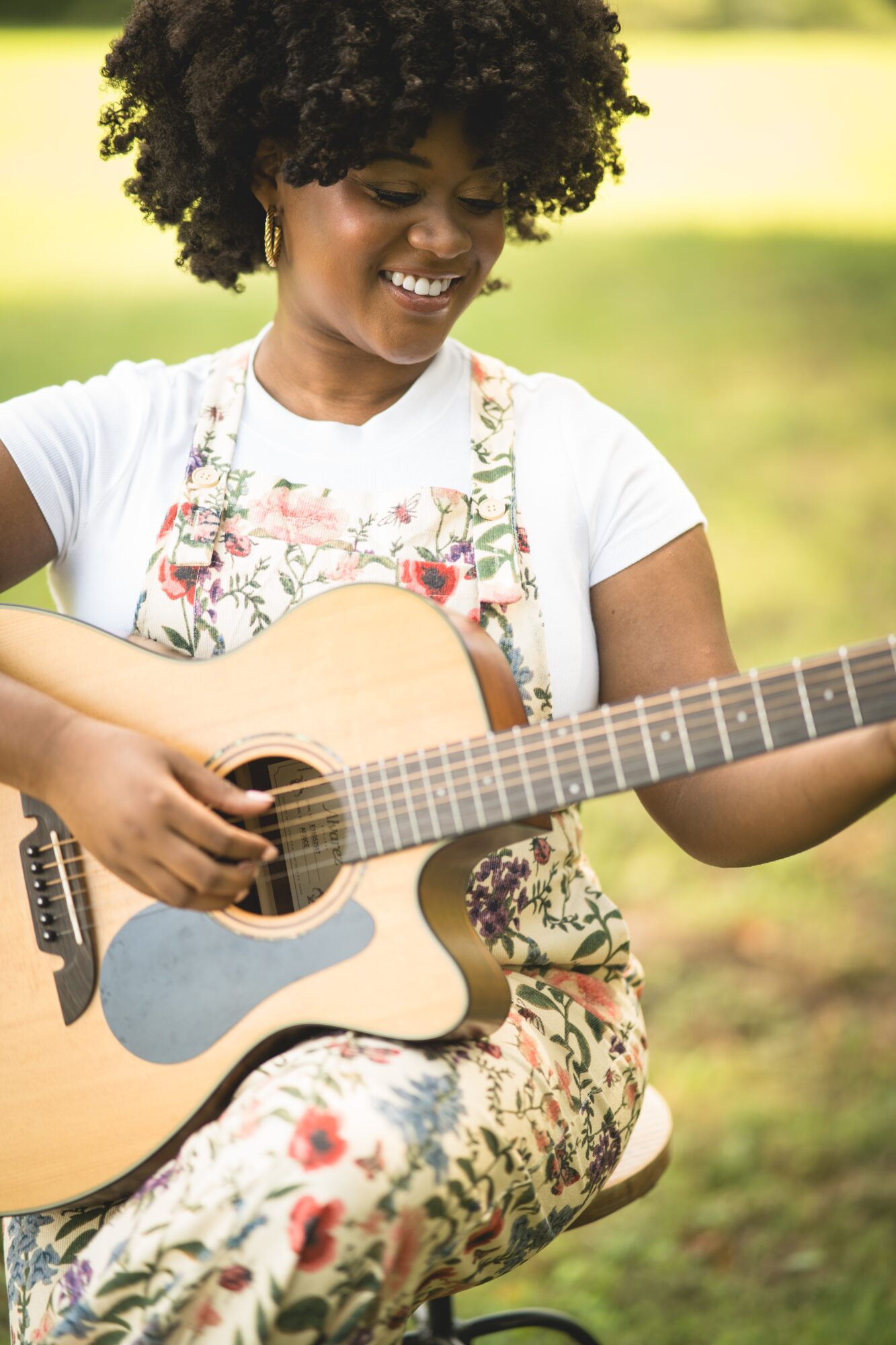 Young woman with curly hair playing an acoustic guitar outdoors, smiling, wearing a floral dress and white top.