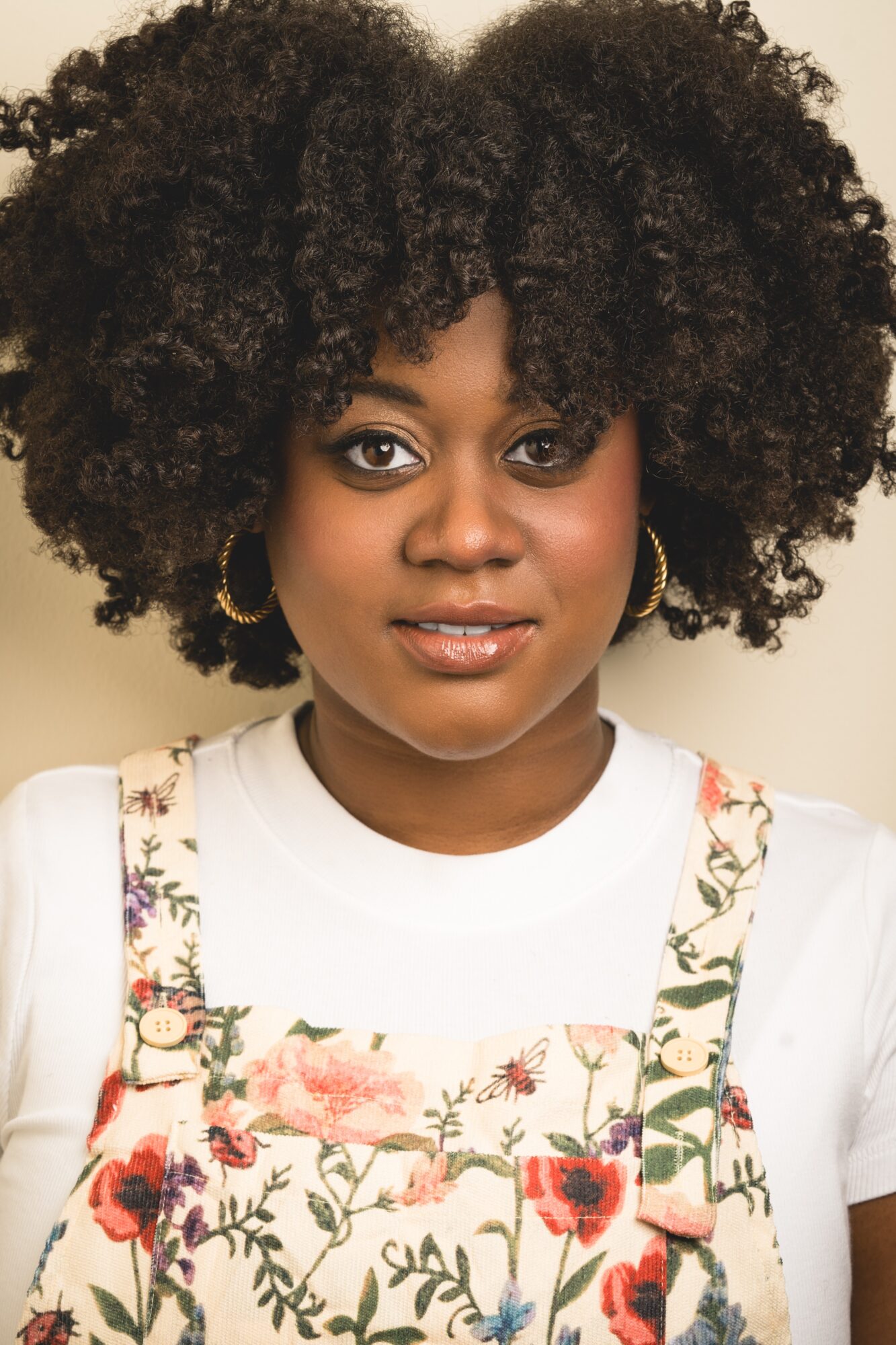Young woman with curly hair, wearing a white shirt and floral overalls, smiling, against a plain background.