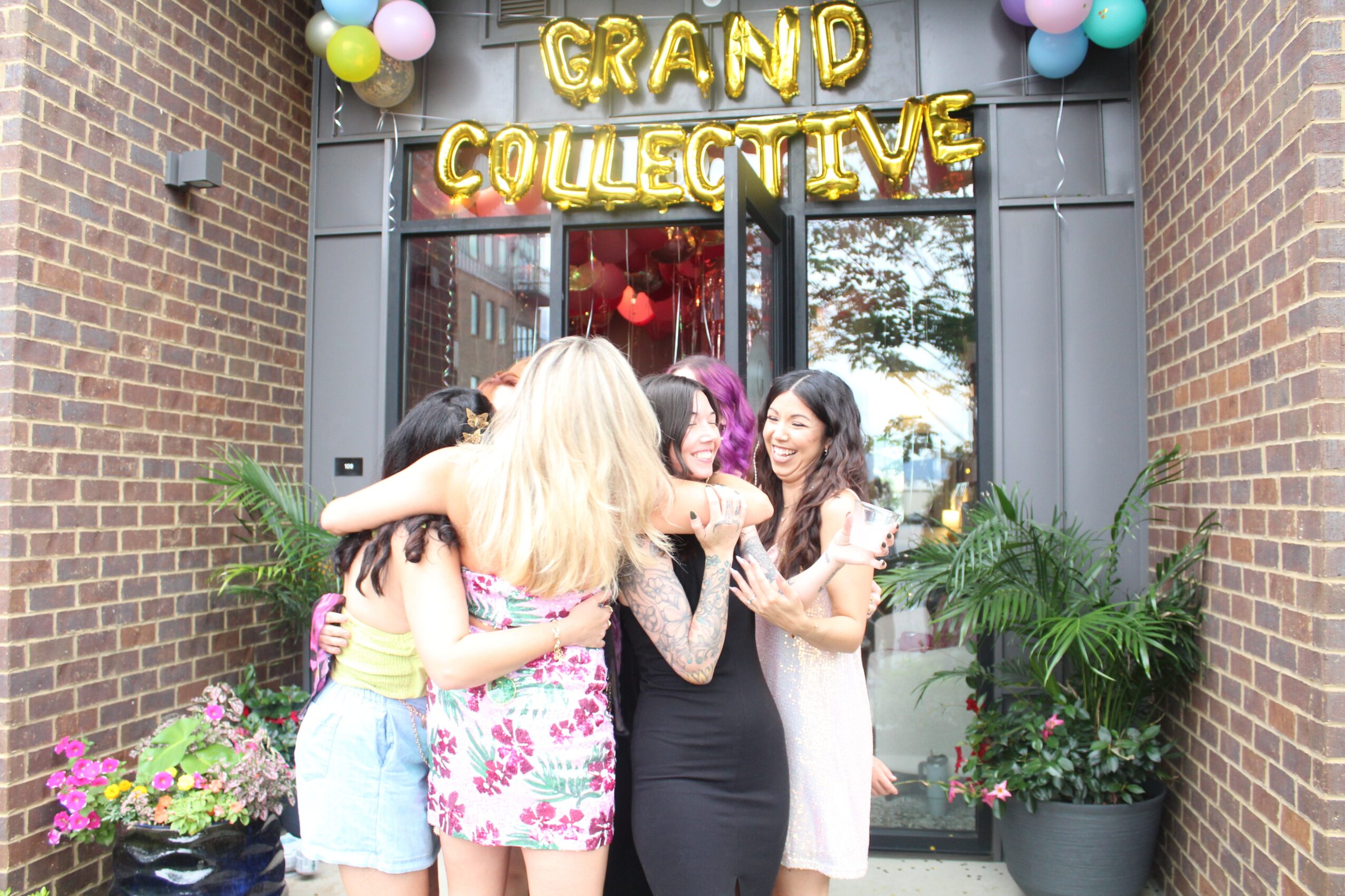 Group of women celebrating in front of a decorated entrance with balloons and a sign reading 'Grand Collective'.