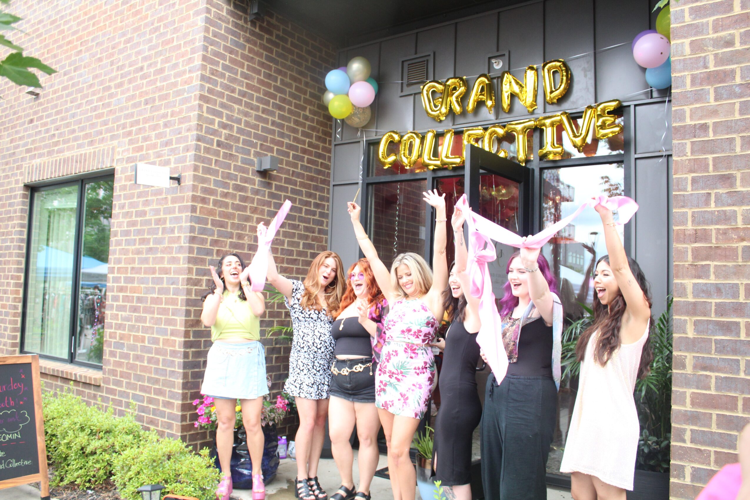 Group of women celebrating outside a building with balloons and a sign that reads 'Grand Collective'.