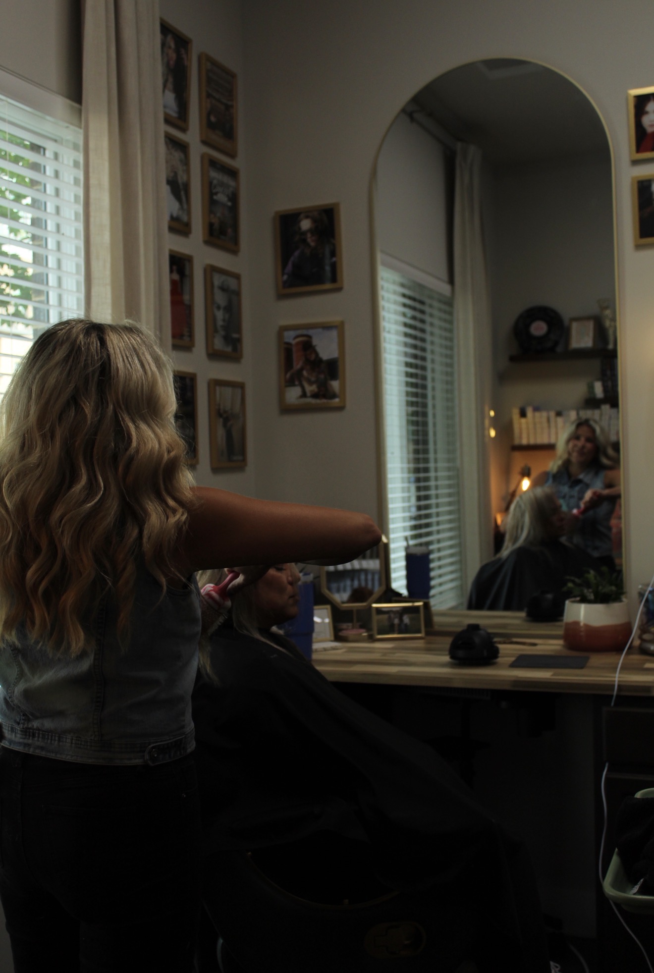 Child with long hair reaching towards a mirror in a room with framed pictures on the wall, window with blinds, and a person sitting in the reflection.