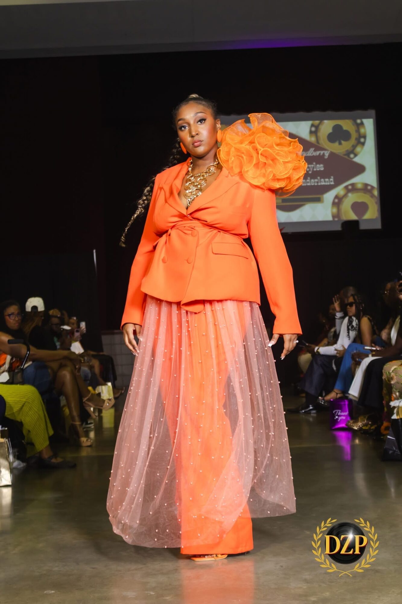 Model walking on runway wearing an orange top with large shoulder details and a pink, glittery, floor-length skirt, audience in background.