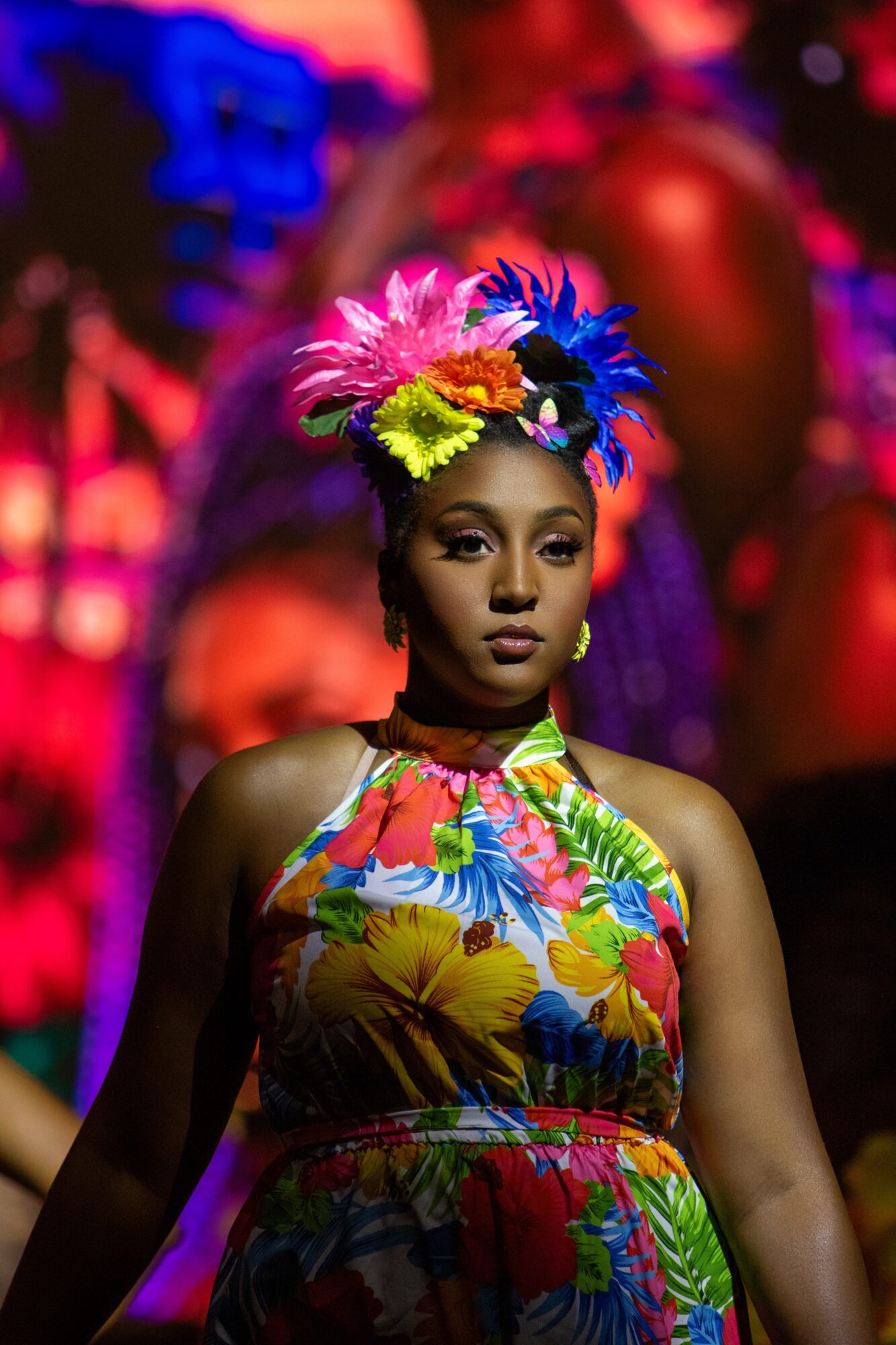 Woman wearing a colorful floral dress and a vibrant flower crown, standing against a blurred colorful background.