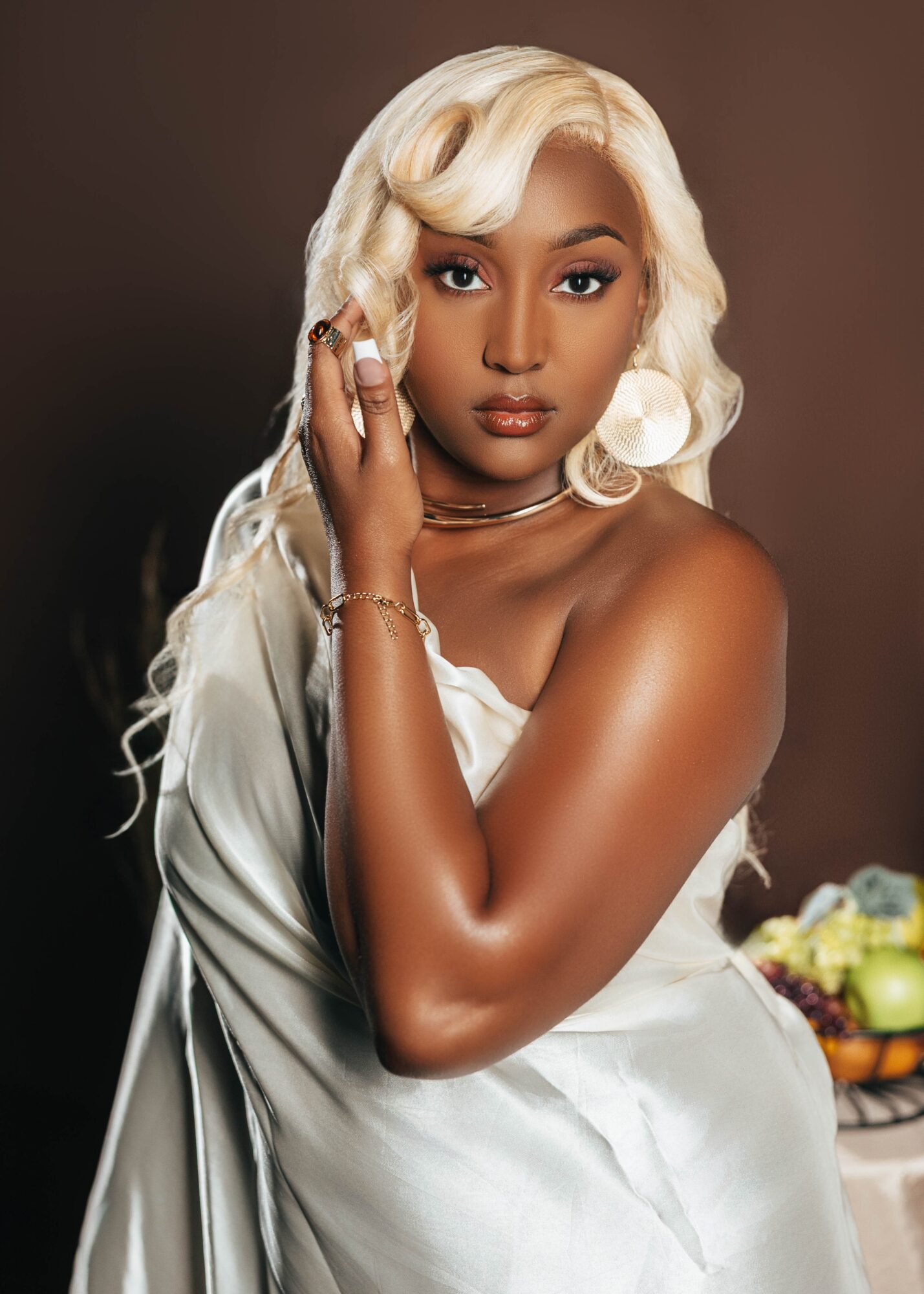 Woman with long, wavy platinum blonde hair, wearing a silky white dress, posing with hand near face, neutral background.