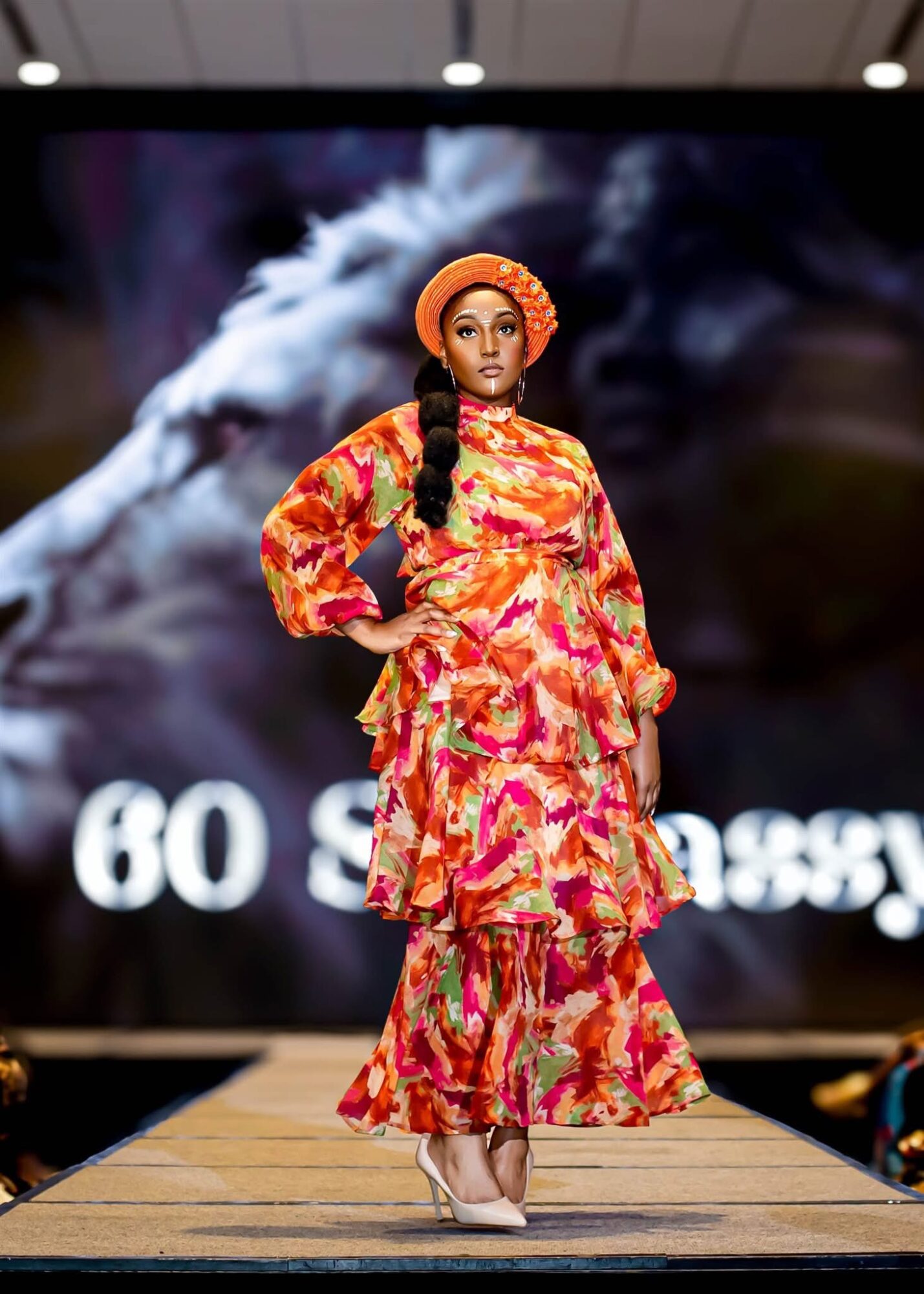Female model walking on runway in colorful patterned dress and orange headwrap, background shows smoky scene and partially visible text.