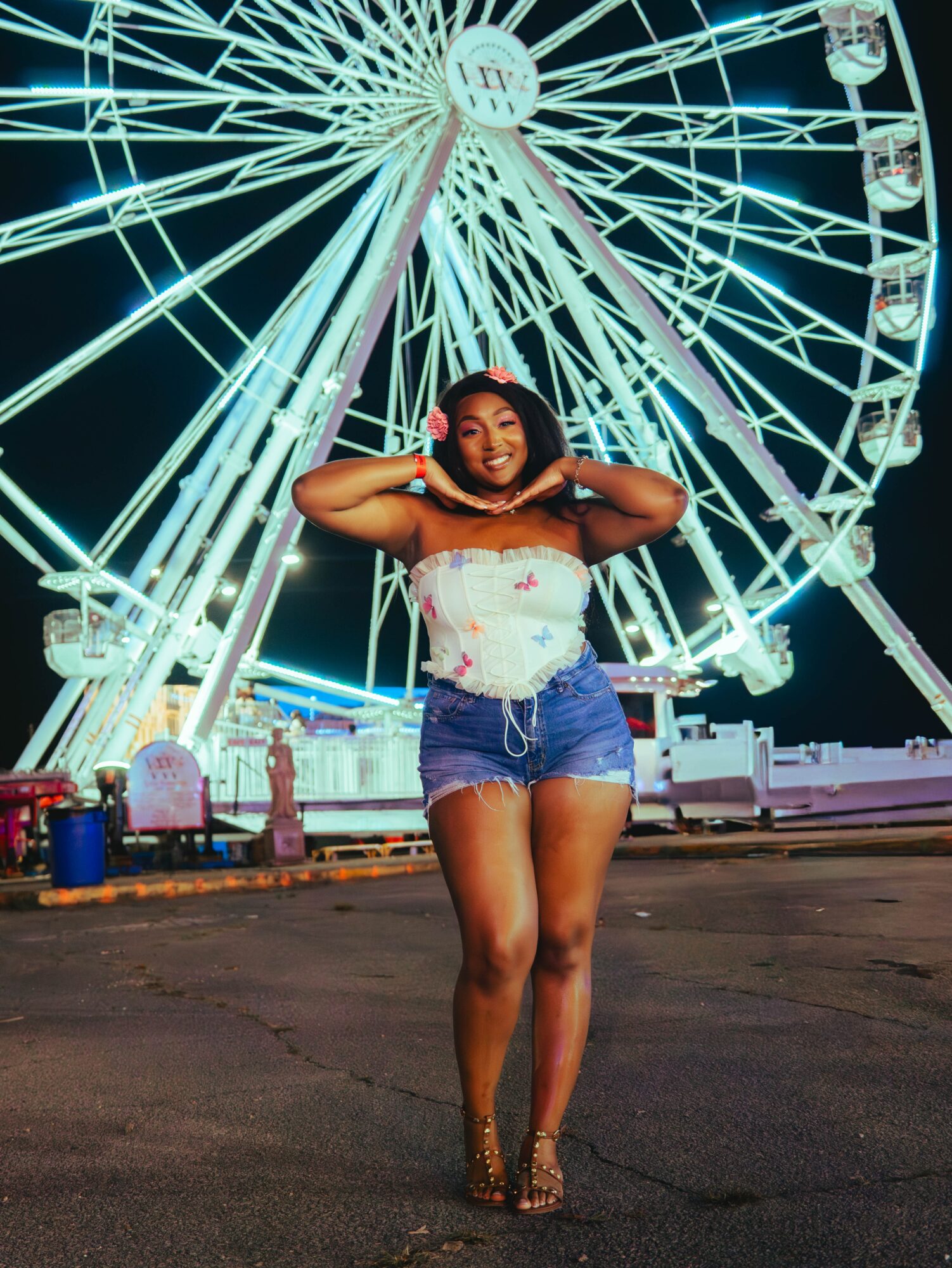 Young woman standing in front of a brightly lit Ferris wheel at night, smiling with hands behind head, wearing a white top and denim shorts.