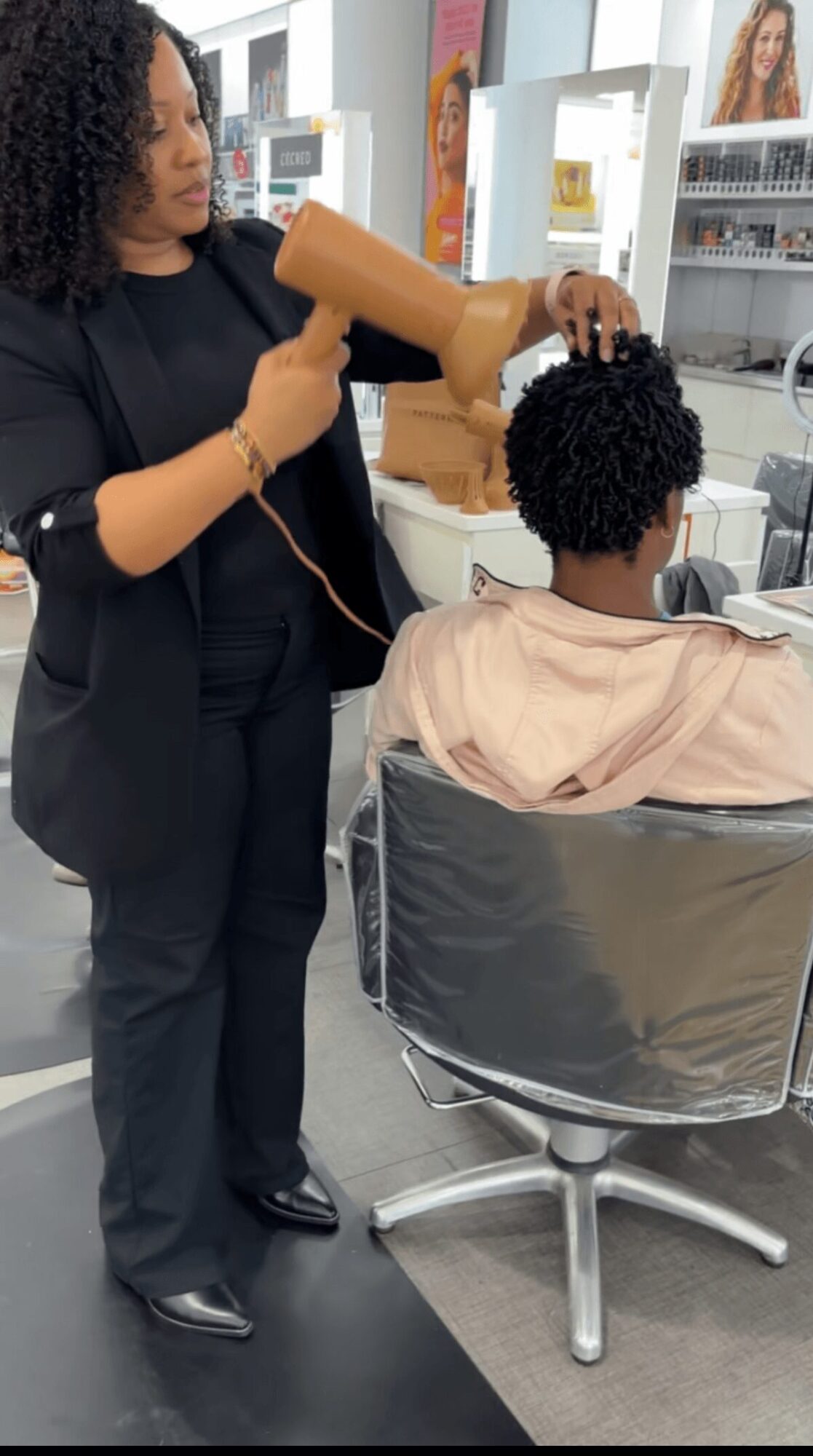 Hairdresser blow-drying a person's curly hair in a salon, with shelves and products in the background.