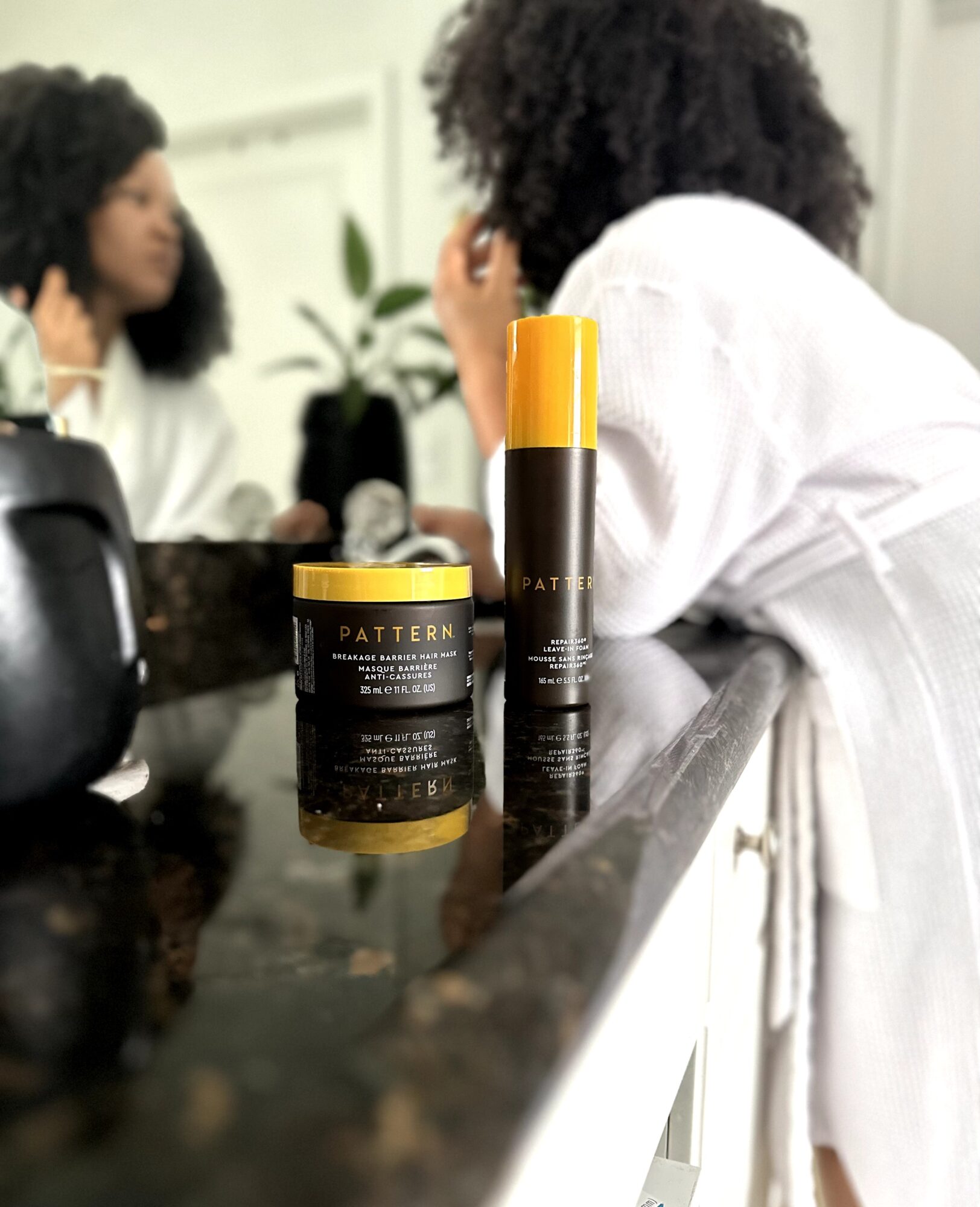 Two women with curly hair sit at a kitchen counter with hair products, a plant, and a black kettle nearby.