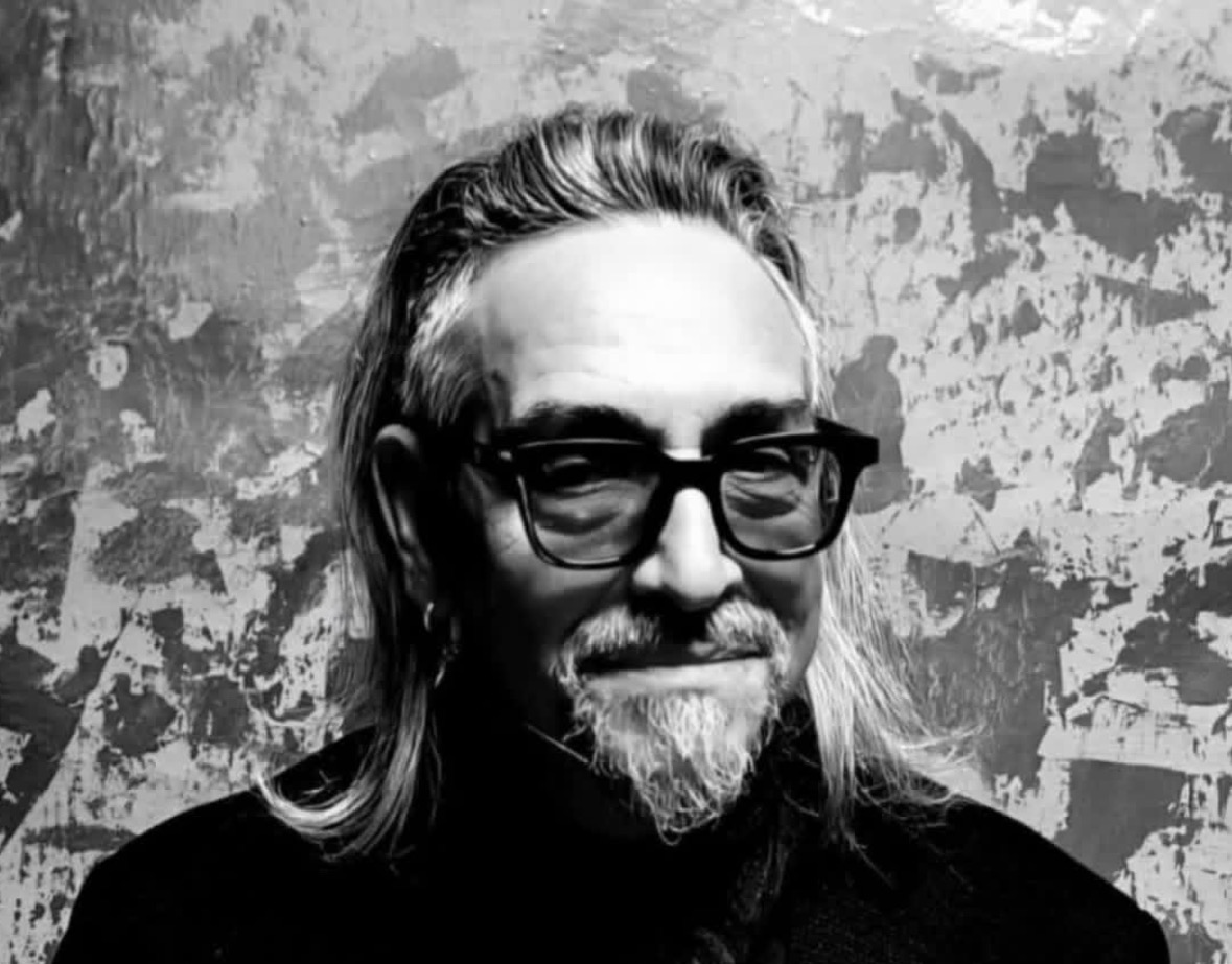 Black and white portrait of a man with glasses, long hair, and a beard, smiling in front of a textured wall.