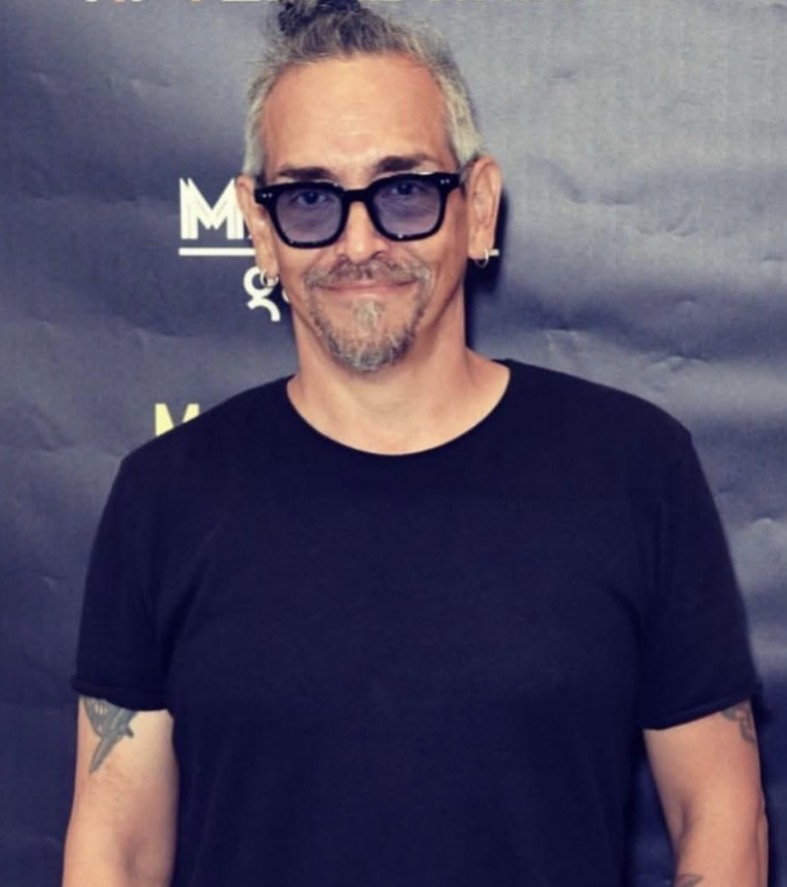 Man with glasses and gray hair, wearing a black t-shirt, smiling, standing in front of a dark background.