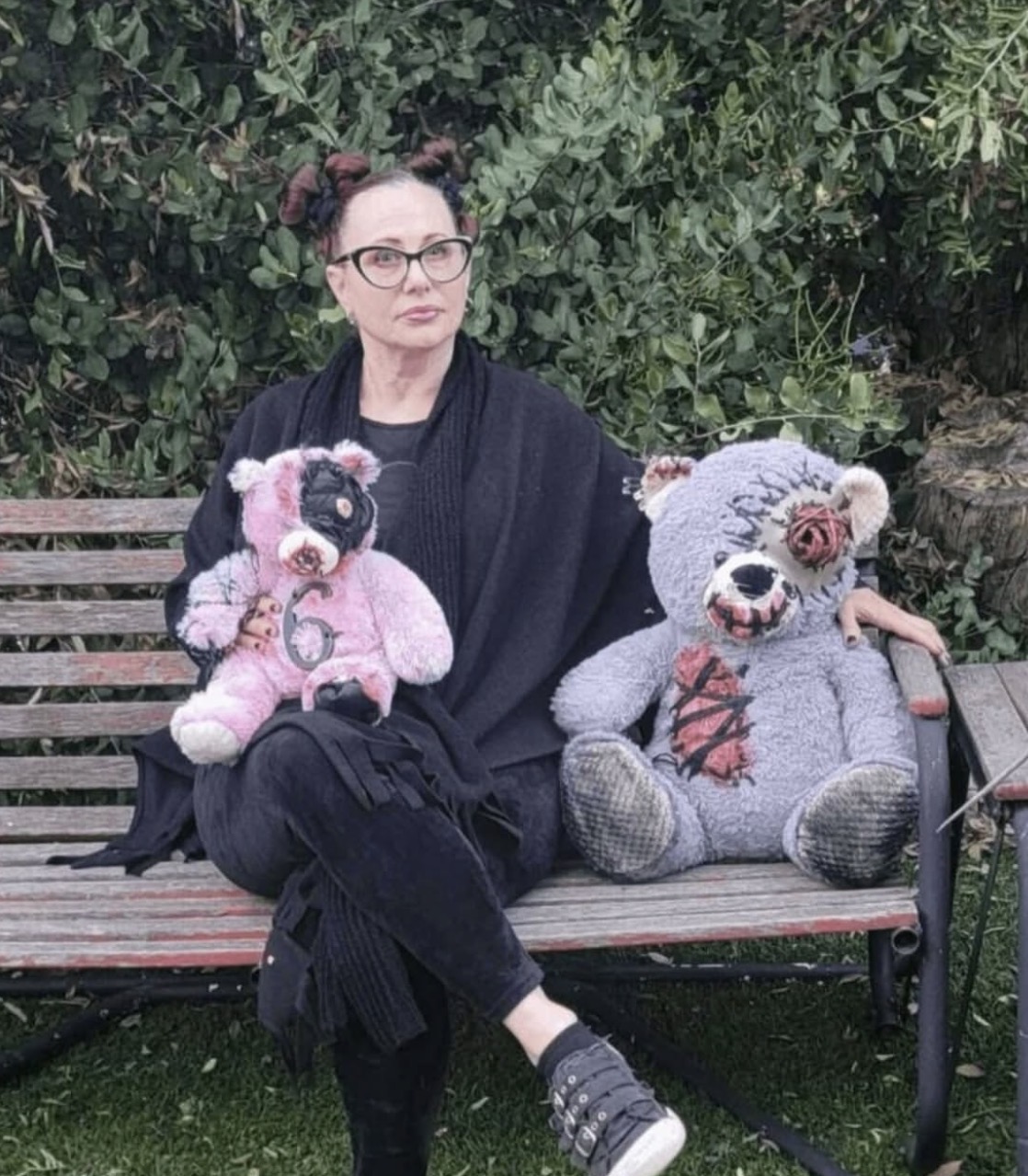 Woman sitting on a park bench holding a pink teddy bear, with a large gray teddy bear beside her, surrounded by greenery.