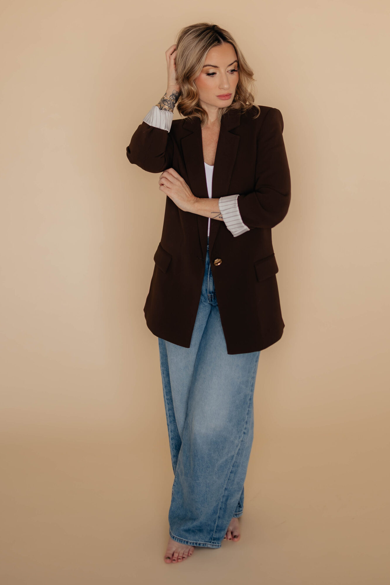Woman with shoulder-length hair in a black blazer, white shirt, and blue jeans, standing against a beige background.