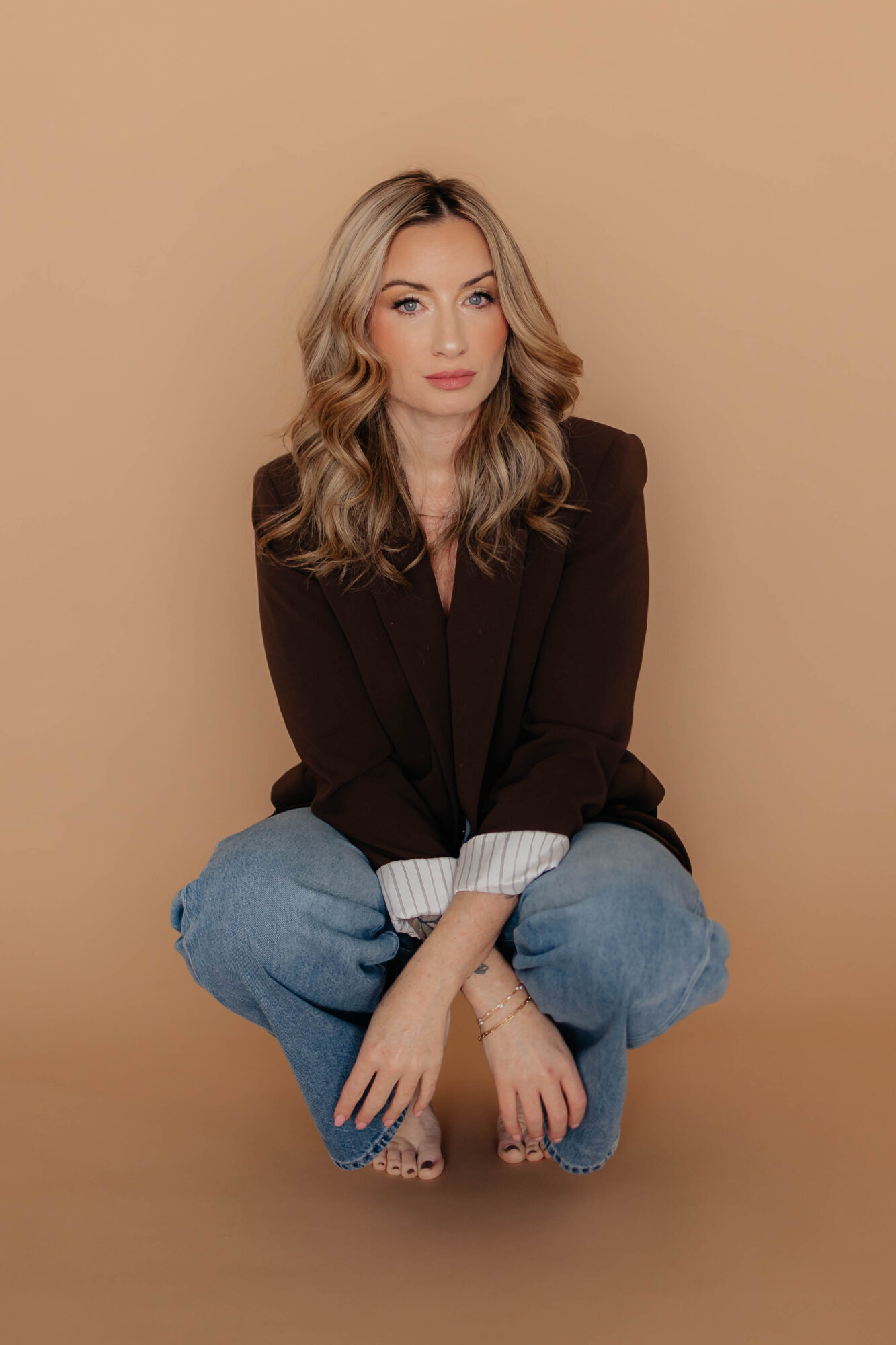 Young woman with wavy blonde hair crouching against a beige background, wearing a dark sweater and blue jeans.