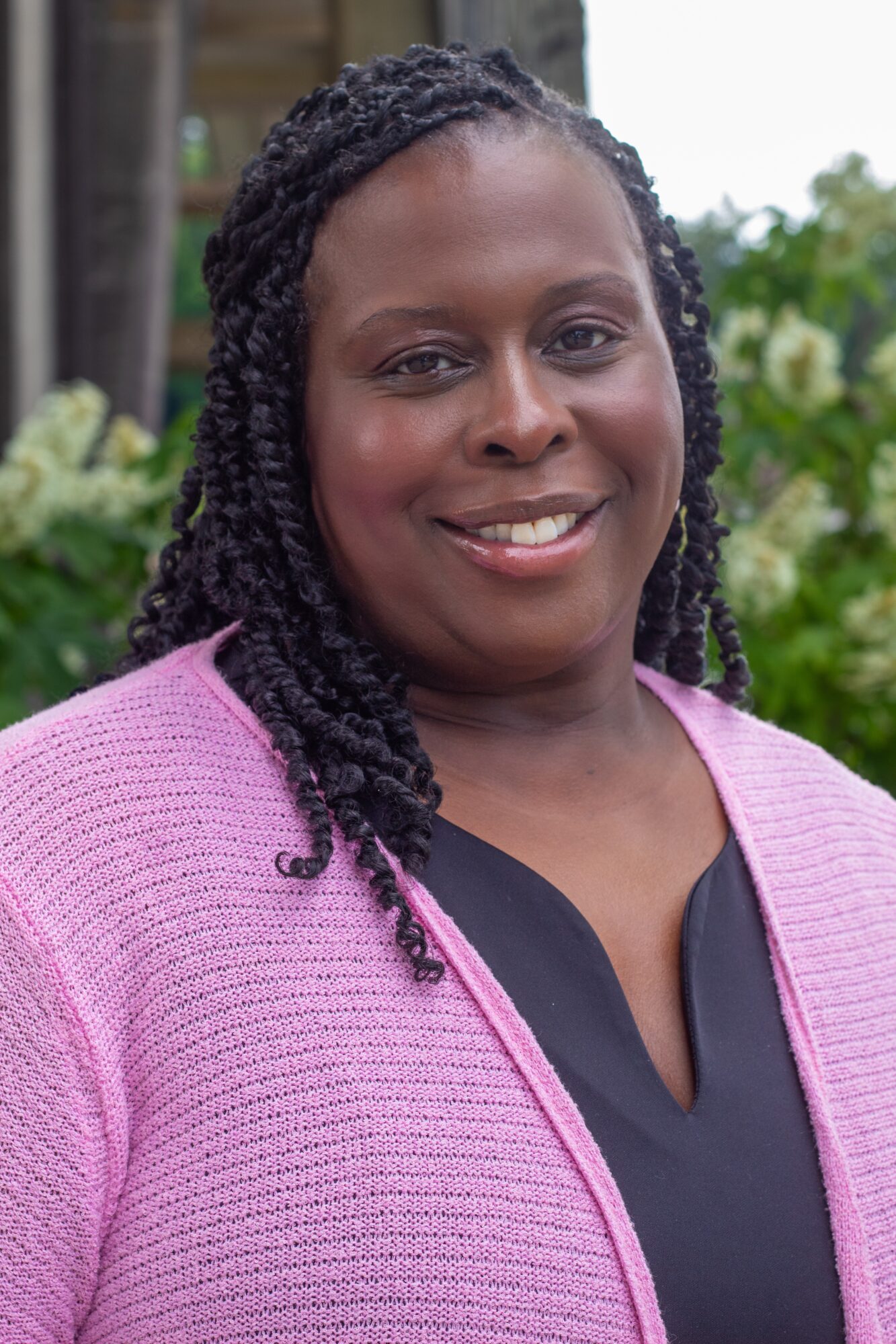 Woman with braided hair smiling outdoors, wearing a pink cardigan over a black top, greenery in background.