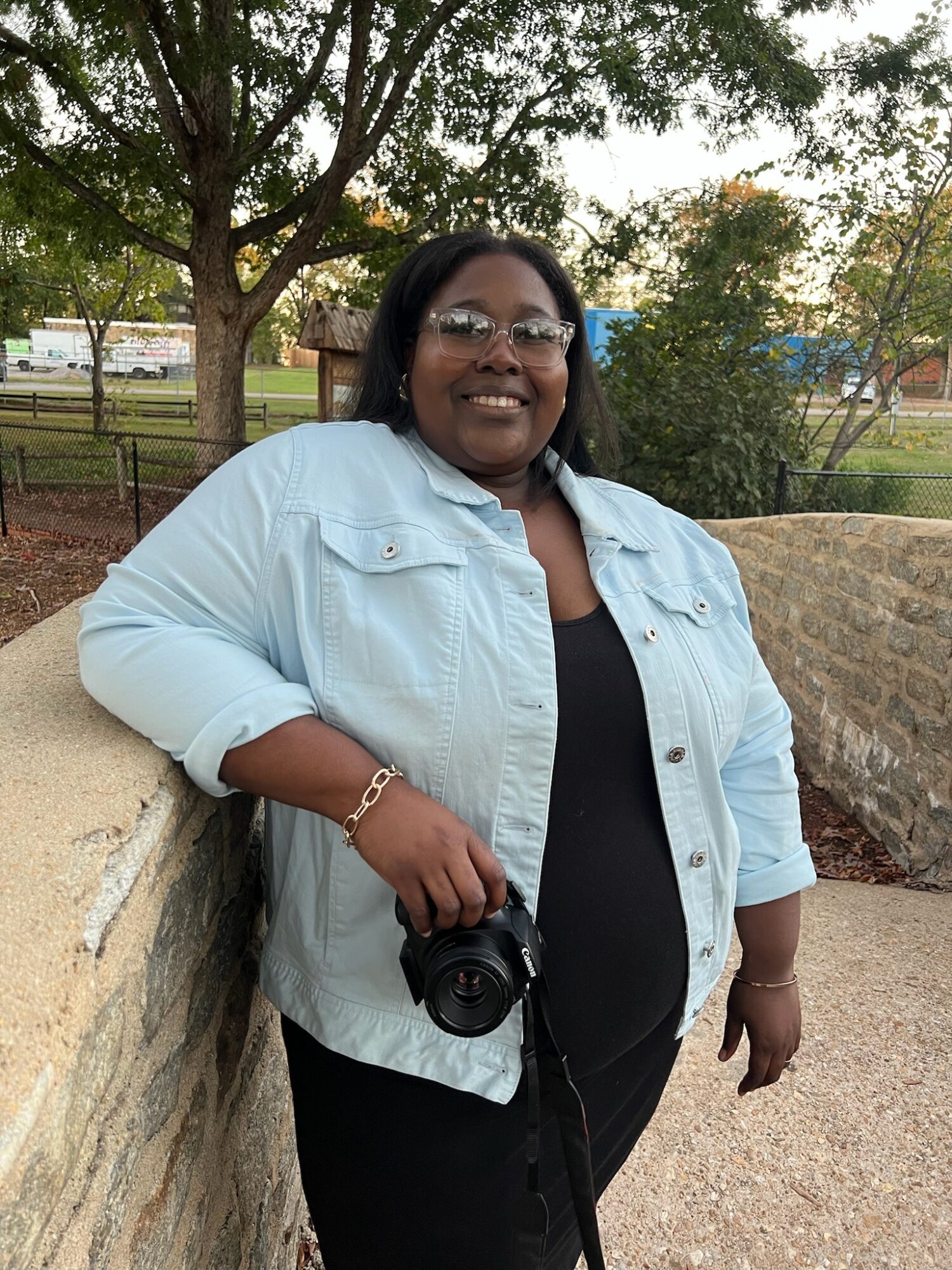 Smiling woman with glasses and a camera, standing outdoors near a stone wall, trees, and a park.