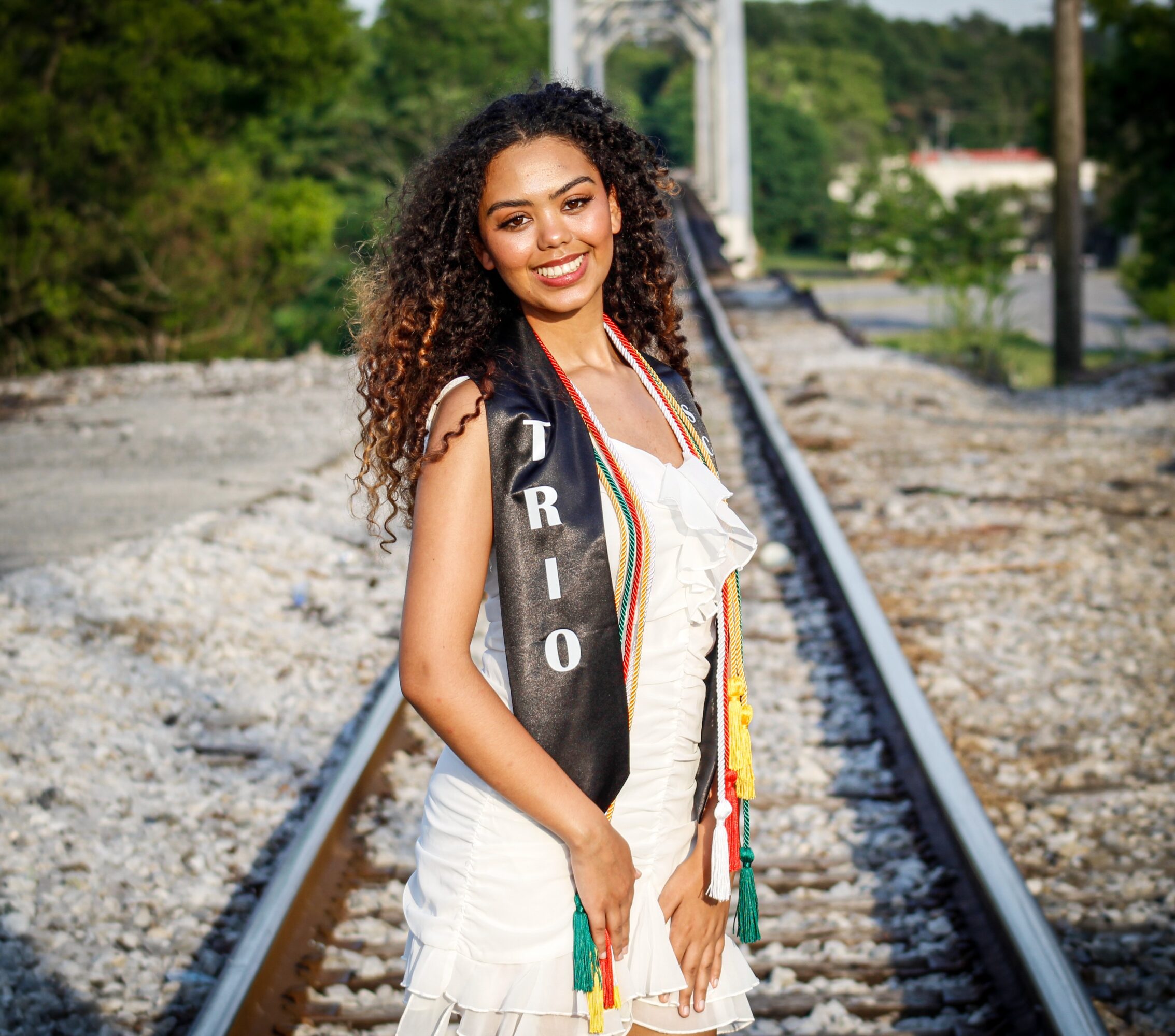Young woman with curly hair standing on train tracks outdoors, smiling, wearing a white dress and a sash with colorful tassels.