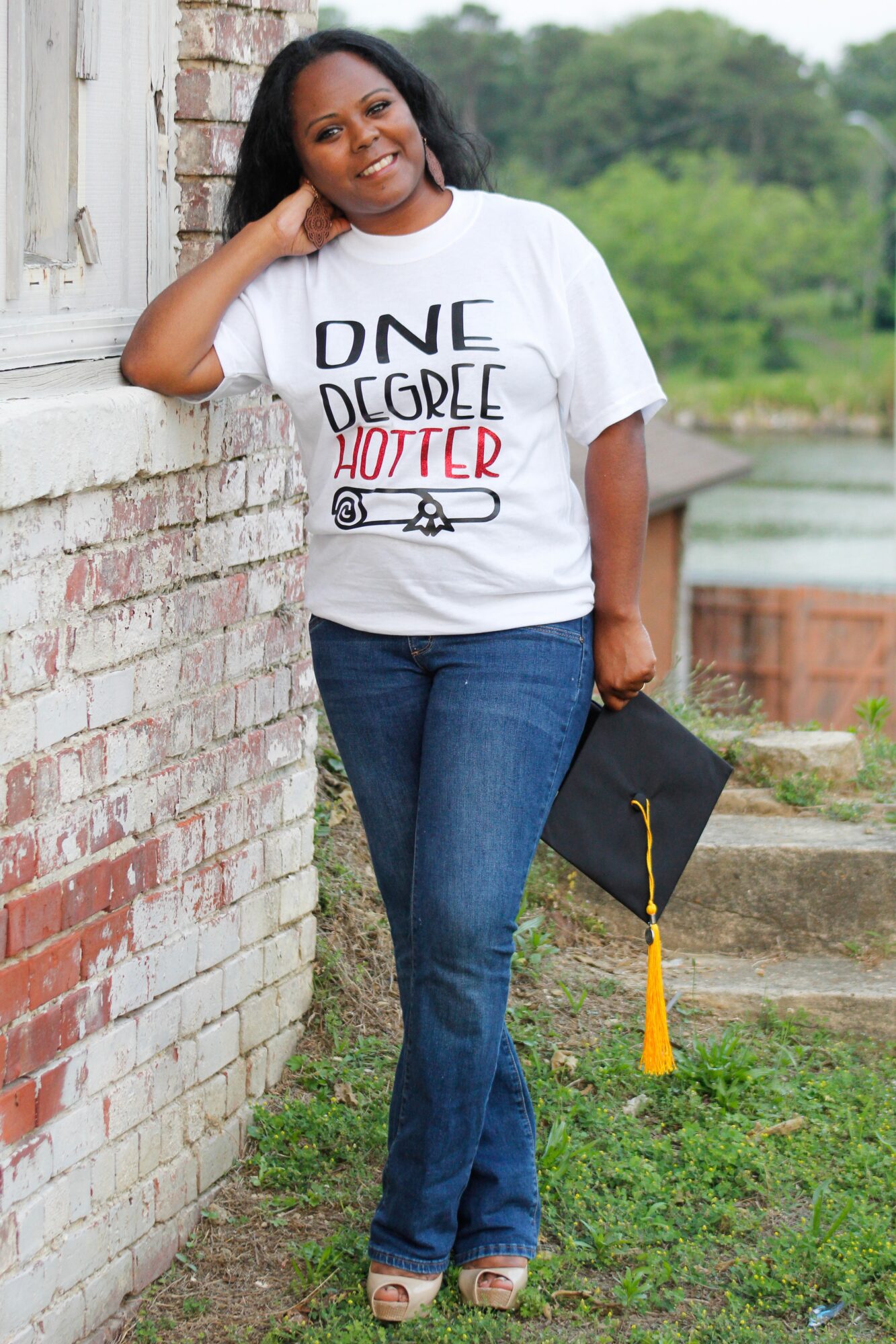 Woman leaning against a brick wall holding a graduation cap outdoors, smiling, with trees in the background.