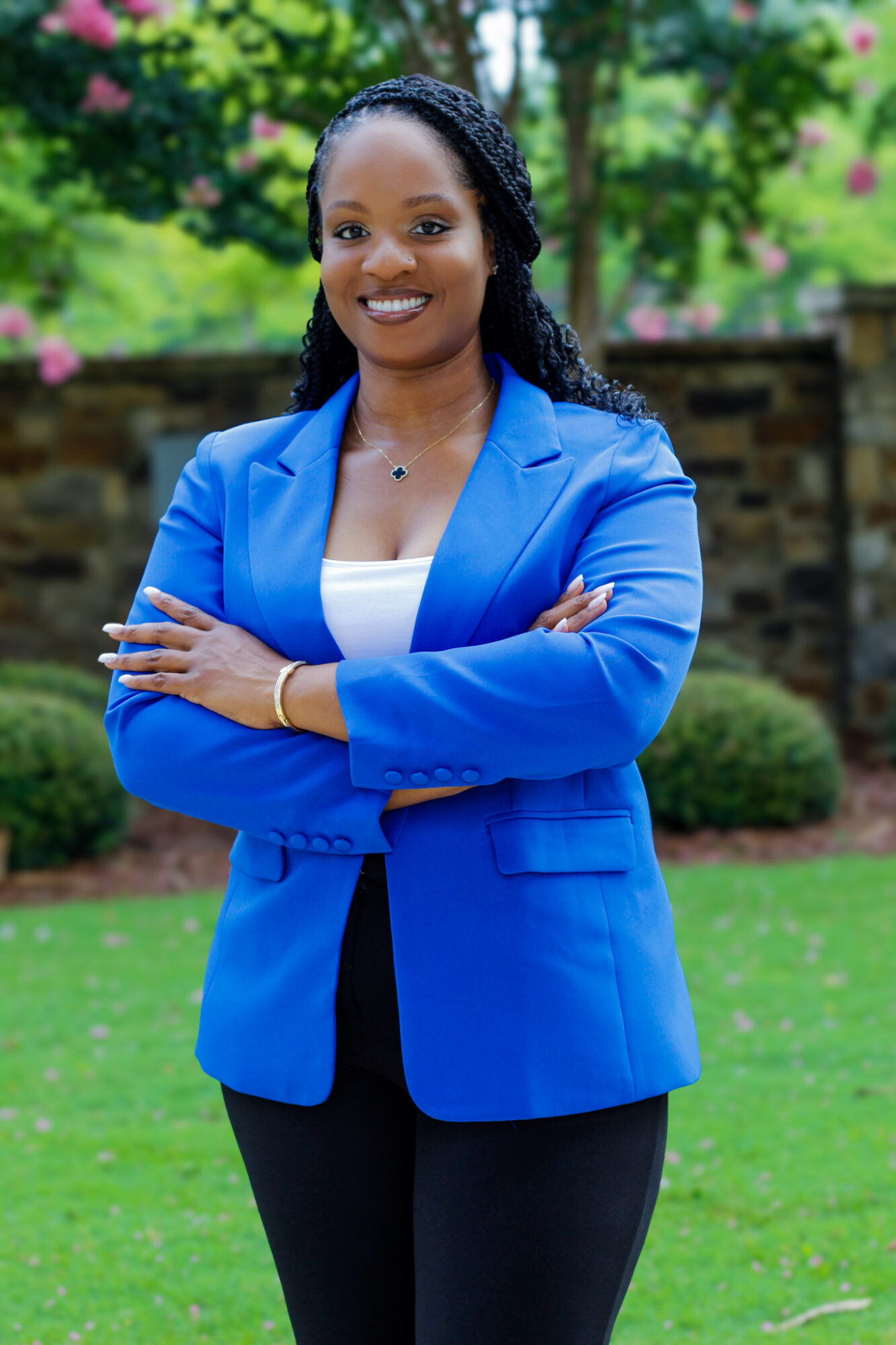 Woman in blue blazer standing outdoors with arms crossed, smiling, greenery and stone wall in background.